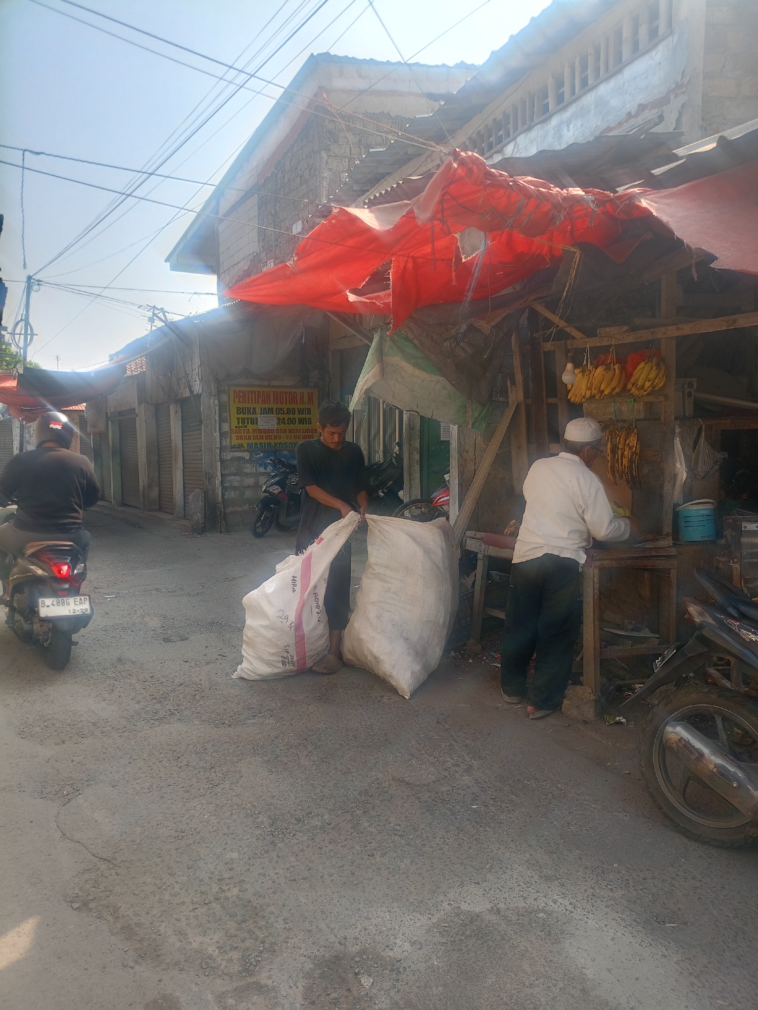 A sunny day scene unfolds in a street market setting in Bojong Gede, Indonesia. In the foreground, a man wearing a black t-shirt and dark pants is handling two large, white sacks, one of which he is opening. To his right, an older man in a white long-sleeved shirt and dark pants, wearing a white cap, is engaged in activity behind a wooden counter at a small stall. Bunches of bananas are hanging from the stall's structure, along with a blue cylindrical container and a light bulb. A motorcycle with the license plate B 4886 EAP is parked and in motion on the left side of the image, with its rider facing away from the camera. Behind the main subjects, buildings with weathered facades and corrugated metal roofs line the street, some covered by torn red tarpaulin. A sign with yellow lettering is visible on a wall to the right, displaying text in Indonesian. The ground is paved with asphalt, showing signs of wear and tear. Several other motorcycles are present in the background. The overall environment appears to be a bustling, informal market area.