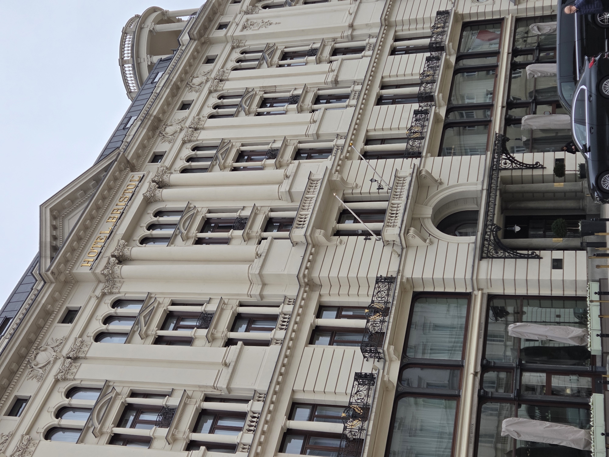 A vertical shot depicts the facade of the Hotel Bristol, located in Warsaw, Poland. The building's architecture features a cream-colored exterior with a series of windows and decorative iron balconies on the right side. The windows appear to have a dark trim and reflect the surrounding environment. The name "HOTEL BRISTOL" is visible in gold lettering on the upper part of the building.
The right side of the image contains a car parked along the side of the hotel. A person is seen standing near the car and appears to be either entering or exiting the vehicle. In front of the car are several table umbrellas. The overall scene suggests a view from ground level, angled upwards to capture the height of the hotel building.