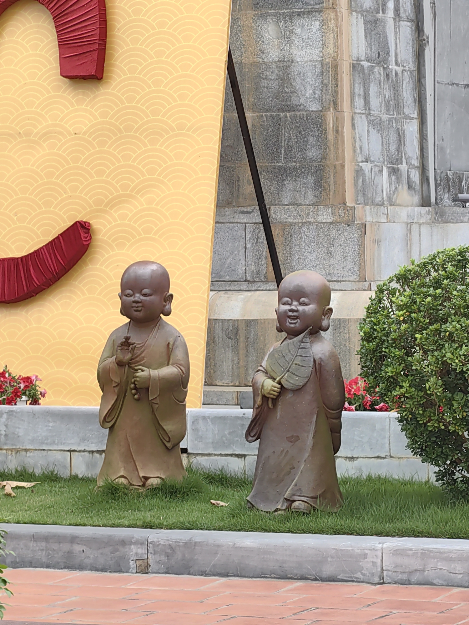Two charming statues of young monks stand side-by-side on a patch of green grass in an outdoor setting, likely a garden or courtyard in Hong Gai Ward, Vietnam.
The monk statue on the left faces forward with a serene expression, its eyes gently closed. It holds a string of prayer beads in its left hand while its right hand is raised in a gesture of blessing or teaching. It wears a simple, flowing robe in a muted earthy brown tone.
The monk statue on the right, also in a similar brown robe, has a wide, joyful smile, with its eyes nearly closed from laughter. It holds a large, textured leaf fan in its left hand, poised as if about to fan itself. Both statues are barefoot.
Behind the statues, a tall yellow wall or display features a repeating, subtle wave-like pattern. Red, pleated fabric decorations, possibly forming parts of a larger symbol, are visible on this yellow backdrop. To the right of the yellow display, a grey stone wall or building with block masonry extends into the background.
The statues are placed on a well-maintained lawn, bordered by a grey concrete curb. Red flowers in pots are visible behind both figures, and a lush green bush partially obscures the background on the right side. The foreground features terracotta-colored pavers, indicating a pathway or patio area.
The scene is brightly lit, suggesting daytime with fair weather. There is no visible text in the image. The overall impression is one of peace, joy, and cultural significance.