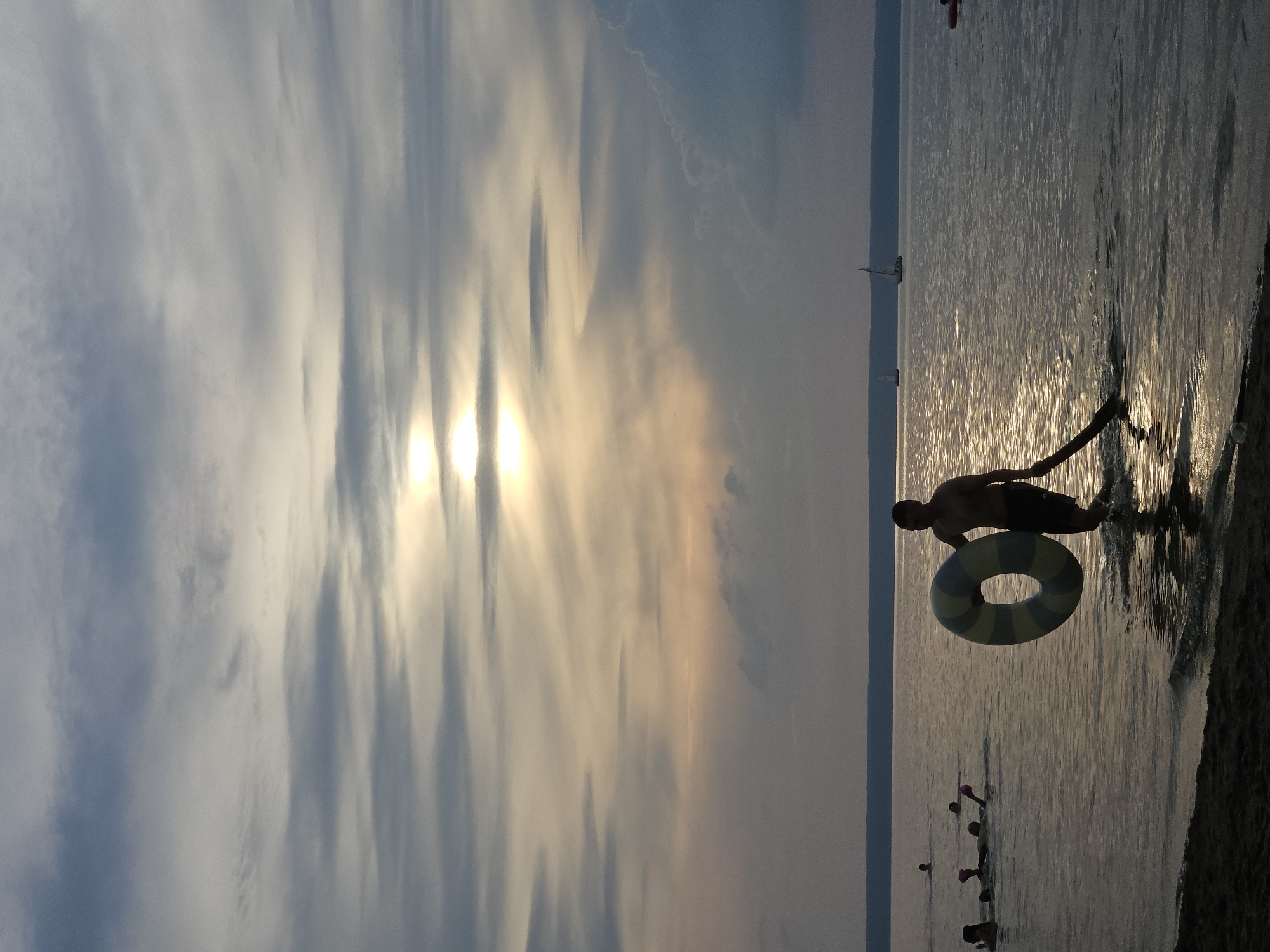 In the city of Leninsky, Russia, a young man walks out of the water at sunset holding a striped inner tube. The sun, partially obscured by clouds, casts a warm glow across the scene. Several other people are visible in the water, some swimming and others appearing to be playing. Two sailboats are in the distance on the calm water. The sky is filled with dramatic, wispy clouds.