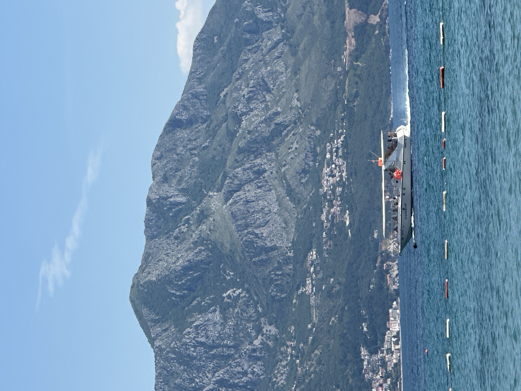 The image captures a scene in Budva, Montenegro. A rugged mountain dominates the left side of the frame, its slopes showing signs of settlement with scattered buildings. The mountain descends to the coastline where more buildings are visible. Adjacent to the coastline is the blue Adriatic Sea, marked by a line of buoys. A speed boat, with the word "SPEEDBOAT" visible on its side, is moving along the water parallel to the coastline.