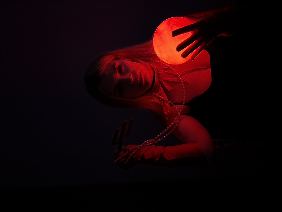 A woman is captured in a dark, red-lit environment, illuminated by a glowing red orb she holds in her hand. She wears gloves and a string of pearls, adding to the scene's enigmatic aura. The composition, with the subject tilted, intensifies the mysterious mood. Her blonde hair and pale skin contrast with the deep shadows, drawing focus to her face and the radiant orb.