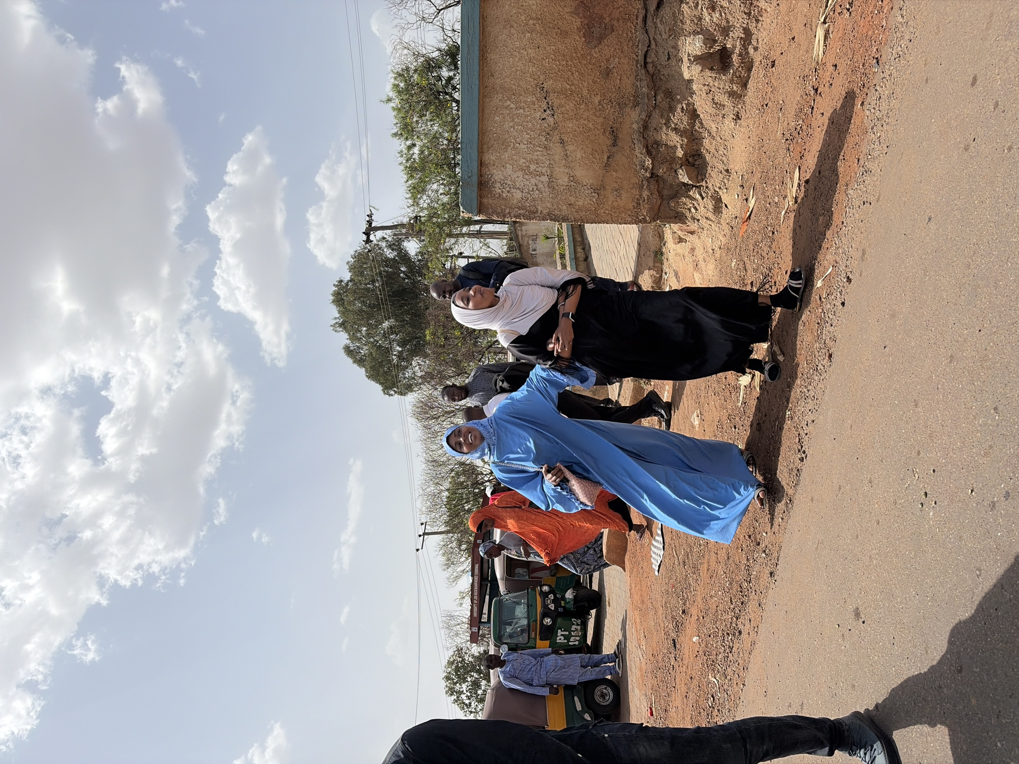 This portrait-oriented photograph captures a sunny street scene in Rafin Par, Nigeria, featuring multiple people, vehicles, and structures under a bright, cloudy sky.
In the mid-ground, two women are prominent. One woman, smiling and looking towards the camera, wears a light blue abaya and a matching light blue hijab. She carries a small, light-colored purse. Beside her, another woman wears a black dress with a white hijab, looking slightly to her left.
Behind these women, several other individuals are visible. One person wears an orange garment, and another wears a patterned shirt. In the background, a green and yellow auto-rickshaw, commonly known as a "keke napep," is parked or moving slowly, bearing the visible text "PT 113ZZ" on its rear. Near the keke napep, a partially obscured sign with red lettering can be seen, possibly including the word "MUNICIPAL."
The setting includes a reddish-brown dirt roadside to the right of the asphalt road, where strong shadows are cast by the midday sun, indicating bright light and clear weather. On the left side, there's a light brown wall with a blue trim, and behind it, a rougher earthen embankment. Utility poles with overhead lines are visible against the sky, and sparse, bare trees suggest a dry season. The sky is a light blue with scattered, fluffy white cumulus clouds.
The scene appears to be a casual gathering or movement of people along a road in a town or semi-urban area, characterized by everyday activity.