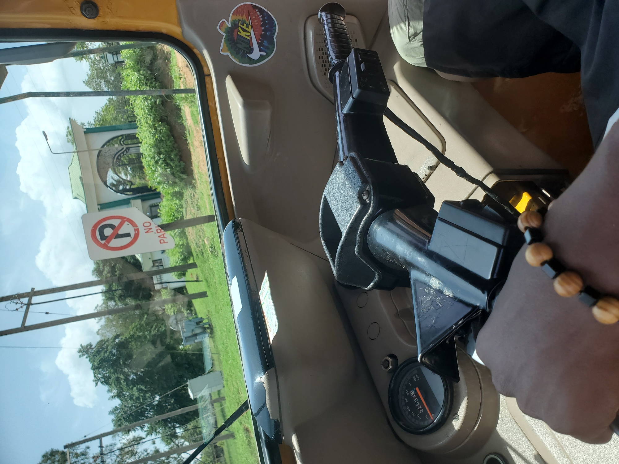 The image provides an interior view from a yellow auto-rickshaw or similar vehicle, looking out through the front window towards a sunny outdoor scene in Vom, Nigeria.
In the foreground, a dark-skinned hand and forearm, adorned with a beaded bracelet featuring light brown/tan and black beads, rests near the vehicle's controls. The bracelet has alternating sets of several light beads and one black bead. Part of the vehicle's black handlebars/steering column is visible, along with a beige dashboard or interior panel. A speedometer gauge is partially visible on the dashboard, displaying "KM/H" and numbers up to 80, with a current reading of "225942" on the odometer. Above the controls, a colorful, tie-dye patterned sticker with the white Nike swoosh and "NIKE" text is affixed to the beige interior trim.
Through the clear window, a bright outdoor scene unfolds under a partly cloudy, blue sky. Prominently in the middle ground is a white building or archway structure with a green, sloping roof and dark arched openings, possibly an entrance gate. Lush green trees and foliage surround this structure and extend across the landscape. In the foreground of the outdoor view, a white "NO PARKING" sign (a red circle with a diagonal line over a black 'P', with "NO PARKI" visible) is partially obscured by the window frame. Several wooden utility poles with wires crisscross the view, suggesting a developed area.
The time of day appears to be bright daylight, indicated by the clear sky and strong shadows. The scene suggests the vehicle is either stationary or moving slowly, providing a clear view of the surrounding environment.