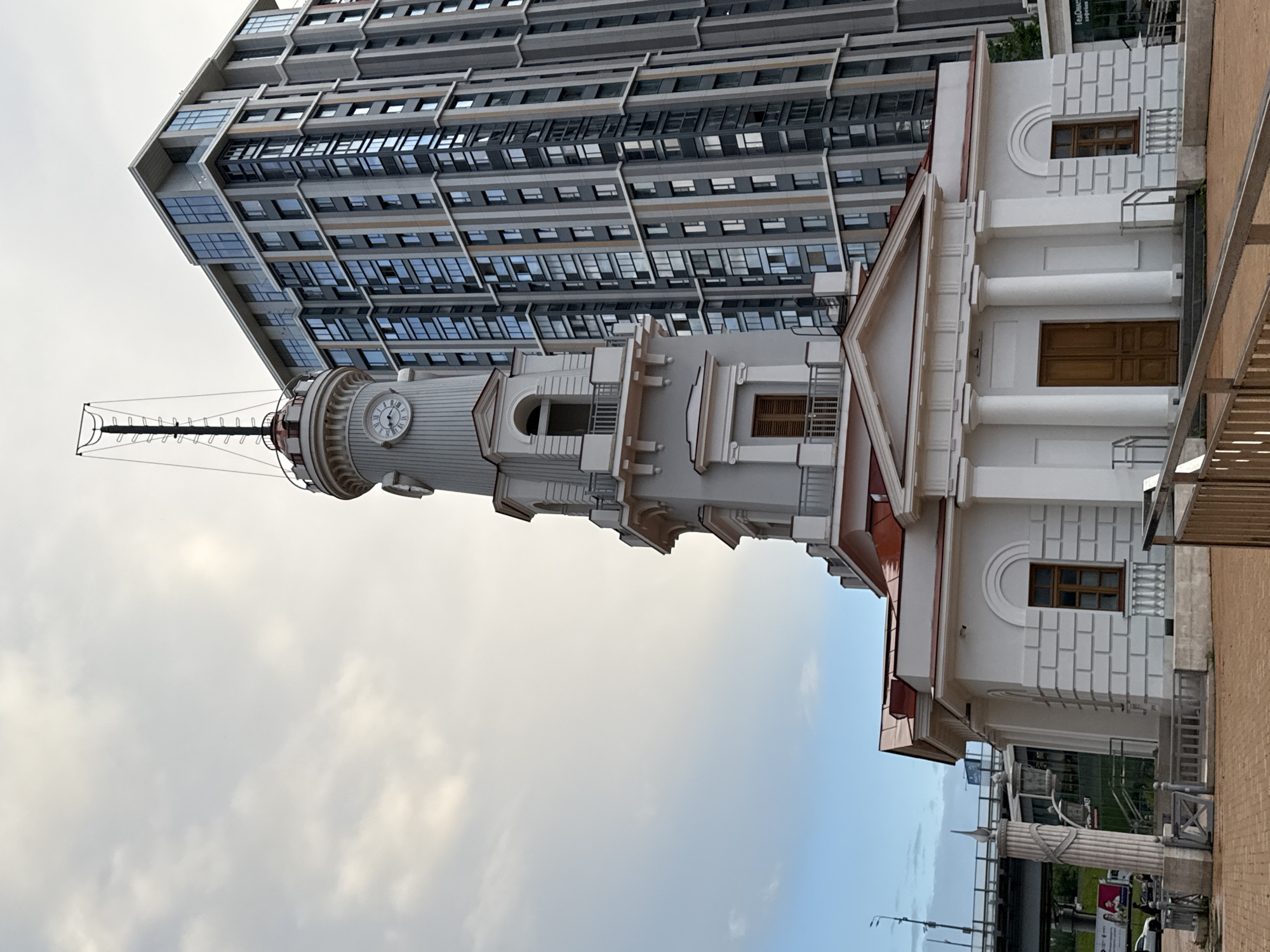 The image shows a historic lighthouse with a clock tower, painted white with a red roof, standing next to a modern, tall, grey apartment building with many windows. The sky above is cloudy and pale. In the foreground, there is a paved area and some metal railings. To the left of the lighthouse, there is a bridge and a street with cars. This is the River Station (Rechnoy Vokzal) in Moscow, Russia, located on the Moscow Canal.