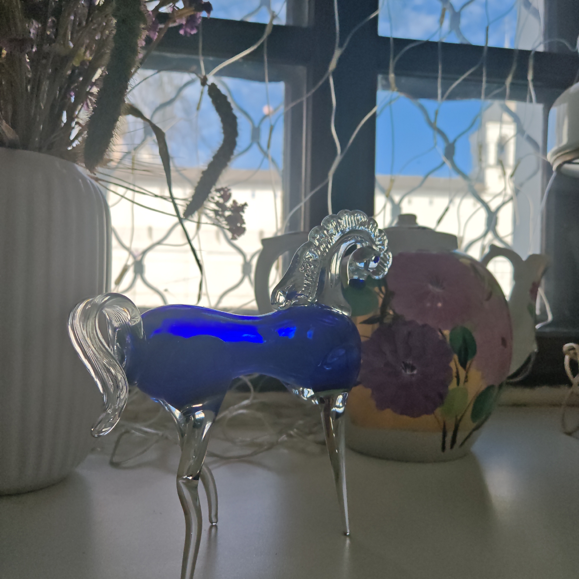 The image features a glass horse figurine, blue in the body and clear in the legs, mane, and tail, placed on a surface near a window. To the left is a white, ribbed vase containing dried flowers. Behind the horse, a floral-patterned teapot is visible. The window, divided into smaller panes with a dark frame, shows a glimpse of a building and a blue sky with clouds. There are also string lights. The scene is staged indoors. There are no people in the image. There is no indication in the image that suggests this is Rostov, Russia.