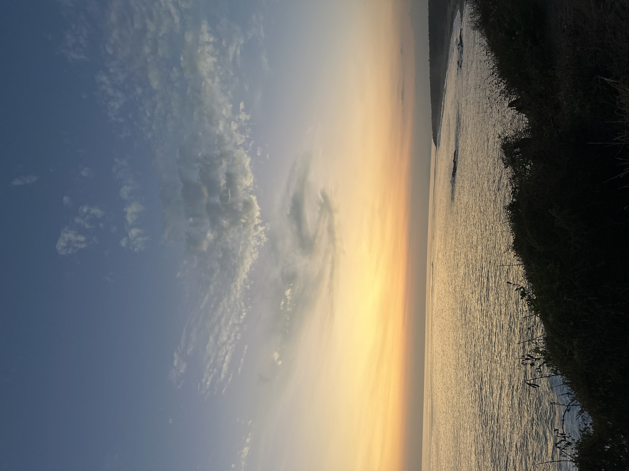 The image captures a coastal sunset scene in Puerto Escondido, Mexico. The sky transitions from blue with scattered clouds to a warm, golden hue near the horizon. The ocean reflects the golden light of the sunset. The right side of the image shows a coastline with vegetation, including grass and bushes. There are no visible people or identifiable subjects in the frame. The focus is on the natural elements of the sky, sea, and coast at sunset.