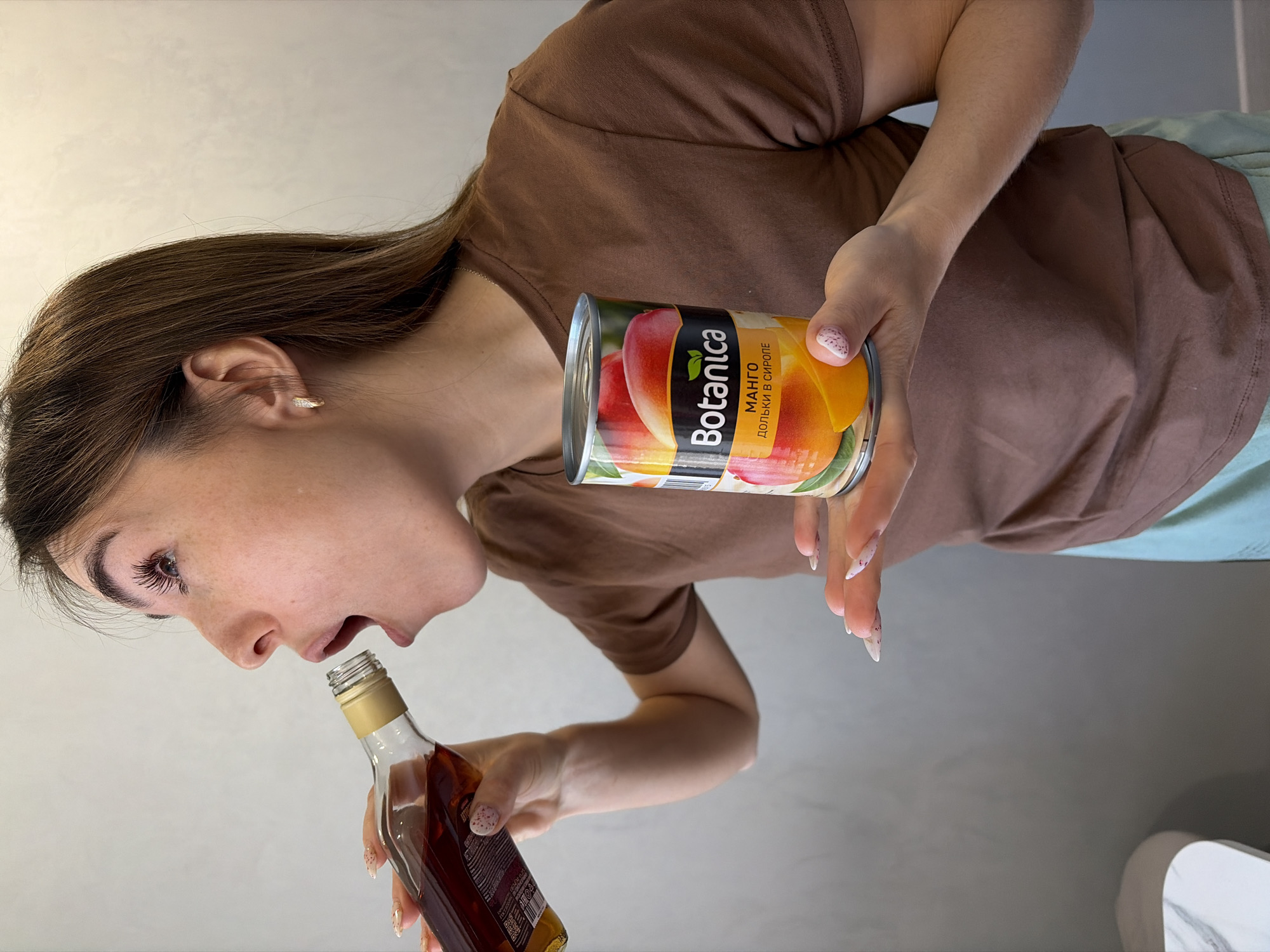 A woman is holding a bottle and a can of "Botanica" brand mango in syrup. The scene appears to be indoors, likely in a home. There is no information to infer any location in the city of Sarapul, Russia.