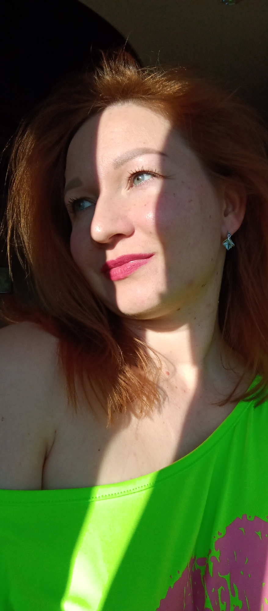 A fair-skinned woman with red hair is captured in a close-up outdoor shot, with strong sunlight casting shadows across her face and neck. She is wearing a bright green top and an earring. Her lips are a shade of pink or magenta. Her gaze is directed to the left. The location is Unknown, Unknown.