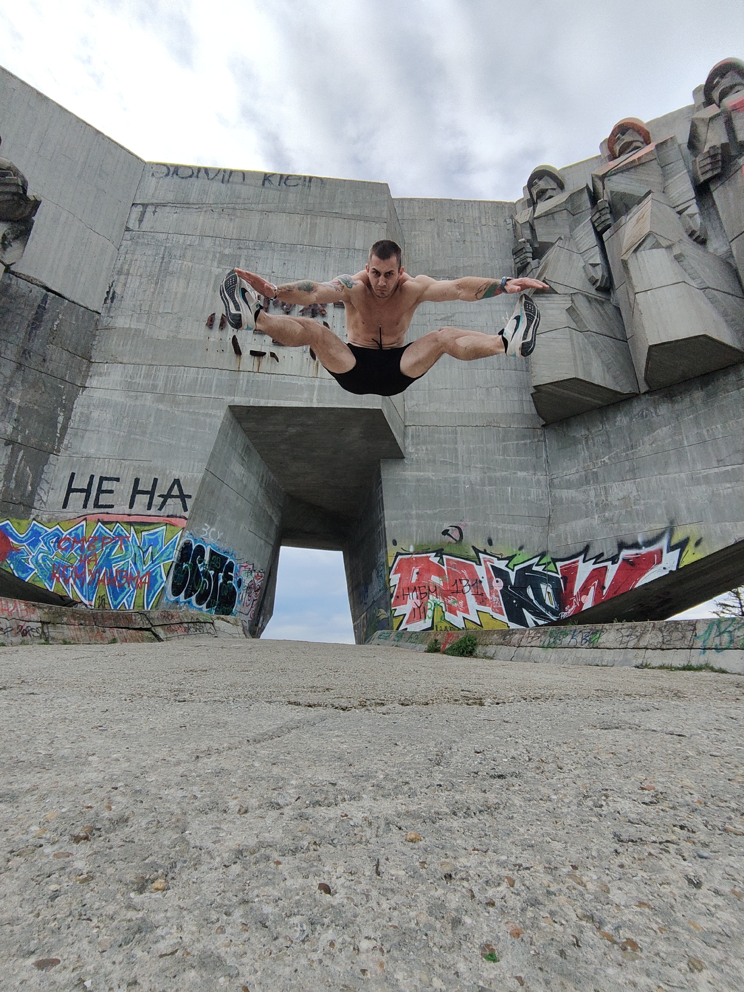 A low-angle, wide shot depicts a shirtless, athletic man mid-air in a split jump, appearing to float against a backdrop of a large concrete structure covered in graffiti, in Buzludzha, Bulgaria. The man is wearing black shorts and sneakers. He is positioned in the center of the frame. His arms are outstretched to the sides, parallel to his legs, indicating balance. The concrete structure behind him features a large opening in the middle, through which a glimpse of the horizon can be seen. The structure is adorned with graffiti, including what appears to be the words "HE HA" in black letters. To the right side of the frame are abstract concrete sculptures. The ground is a rough, weathered concrete surface, and the sky is overcast.