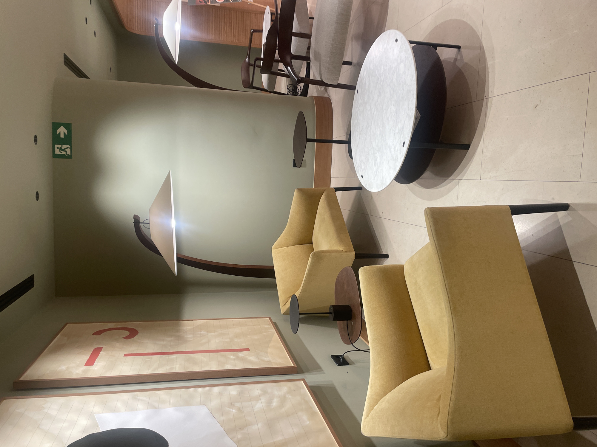 The image depicts an indoor seating area, likely a lounge or waiting area, with a modern and minimalist aesthetic. Two yellow upholstered armchairs are prominently featured in the foreground, one facing left and the other to the right, angled slightly towards the center. Between these chairs and slightly further back, a small, dark-colored side table with a cylindrical lamp sits on a dark brown, oval-shaped tabletop. To the right of the yellow chairs, a larger, oval-shaped coffee table with a white marble-patterned top rests on dark legs, with a dark wood lower shelf. Behind the coffee table, two wooden chairs with light-colored upholstered seats and backs are visible, appearing to be more formal seating.
The walls are painted a muted olive green. Two large abstract artworks hang on the left wall. The artwork on the left has a large black circle and white geometric shapes. The artwork on the right features vertical red lines and a red curved shape on a light background. Above the seating area, an arched floor lamp with a cone-shaped shade is positioned behind the yellow chairs. In the upper right portion of the image, a green exit sign with a running figure and an arrow is visible on the curved wall, indicating a potential emergency exit. The flooring consists of large, light-colored square tiles. The lighting appears to be a combination of ambient room lighting and the lamps, creating soft shadows.
The scene does not contain any people or indicate any specific actions or interactions. The environment suggests a public or semi-public space designed for relaxation or waiting. The presence of the exit sign implies it is an indoor public building. The overall impression is one of contemporary design and comfort. There is no visual information in the image that links it to the city of Sevilla, Spain.