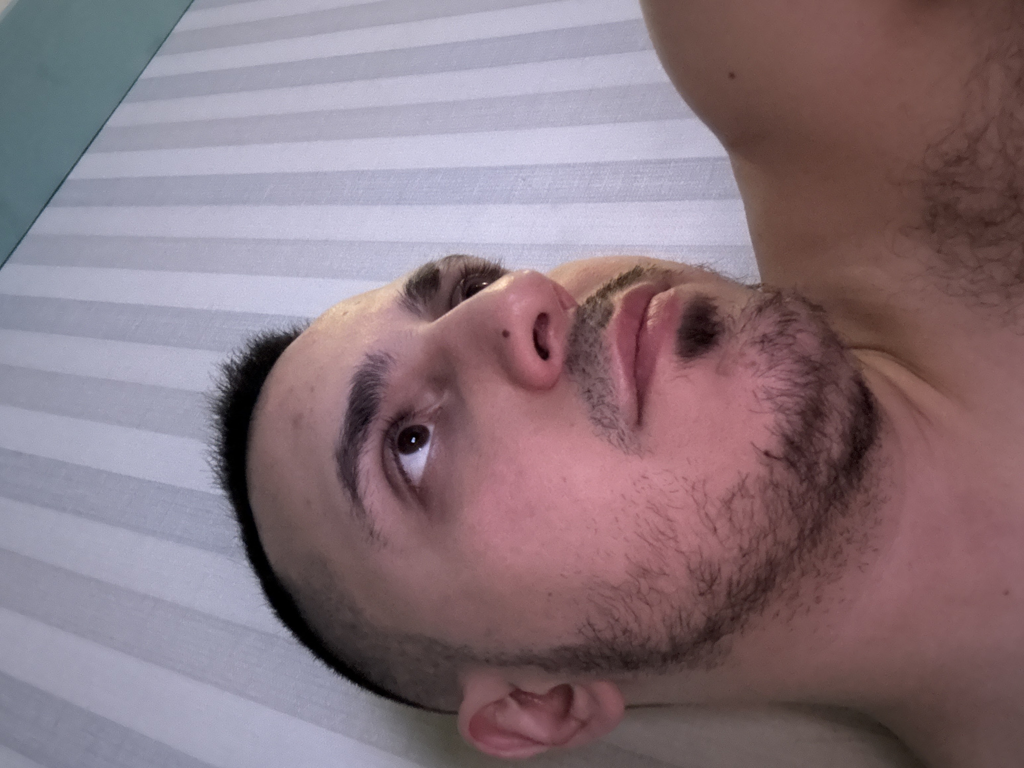 The image features a close-up, high-angle shot of a man lying down, looking upwards. He is positioned horizontally in the frame, with his head on the left and body extending to the right. The man has short, dark hair, dark eyebrows, and a short beard or stubble. His gaze is directed upwards and slightly to his left, with his right eye visible and focused. His skin appears fair to medium-toned. Part of his upper body, including a shoulder and chest area with some body hair, is visible on the right side of the frame, suggesting he is shirtless or partially clothed.
The background consists of a surface with horizontal stripes of alternating light gray and white or very light blue. This patterned surface covers most of the background, likely a bed sheet or wallpaper. In the top left corner, a solid light blue or teal colored surface is visible, possibly another part of a bed or furniture. The lighting suggests an indoor setting, but the specific time of day is not discernible. No text is visible in the image. The man's expression appears calm and contemplative.