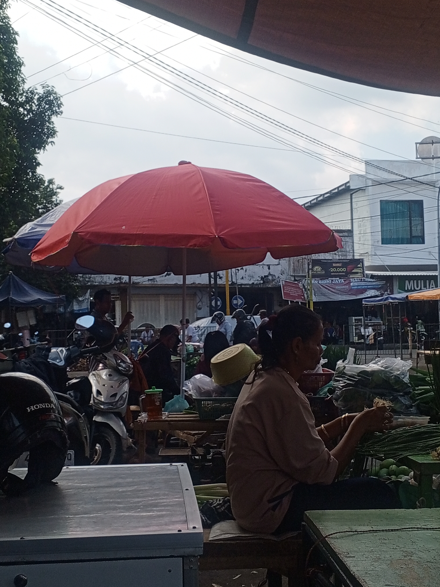 This is a photograph of a bustling street market scene in Jember, Indonesia. The foreground is dominated by a portion of a silver food cart or stall. To the right of the cart, a woman is seated, facing away from the camera, wearing a light brown shirt and dark pants. She is actively engaged in preparing some sort of food item, holding strands of what appears to be noodles or vegetables in her hands. Her hands are adorned with bracelets. Behind her, a variety of produce is neatly arranged, including bundles of green vegetables and round, light green fruits, possibly limes or small gourds. To the left and behind the woman, a large red umbrella provides shade, and beneath it, several motorcycles are parked. In the middle ground, a man in a dark shirt is visible, appearing to be tending to his stall. Further in the background, other market stalls, awnings, and a white building with a prominent green window are discernible. The sky is overcast with visible power lines stretching across the frame. The overall impression is that of a lively market day with vendors and shoppers interacting.