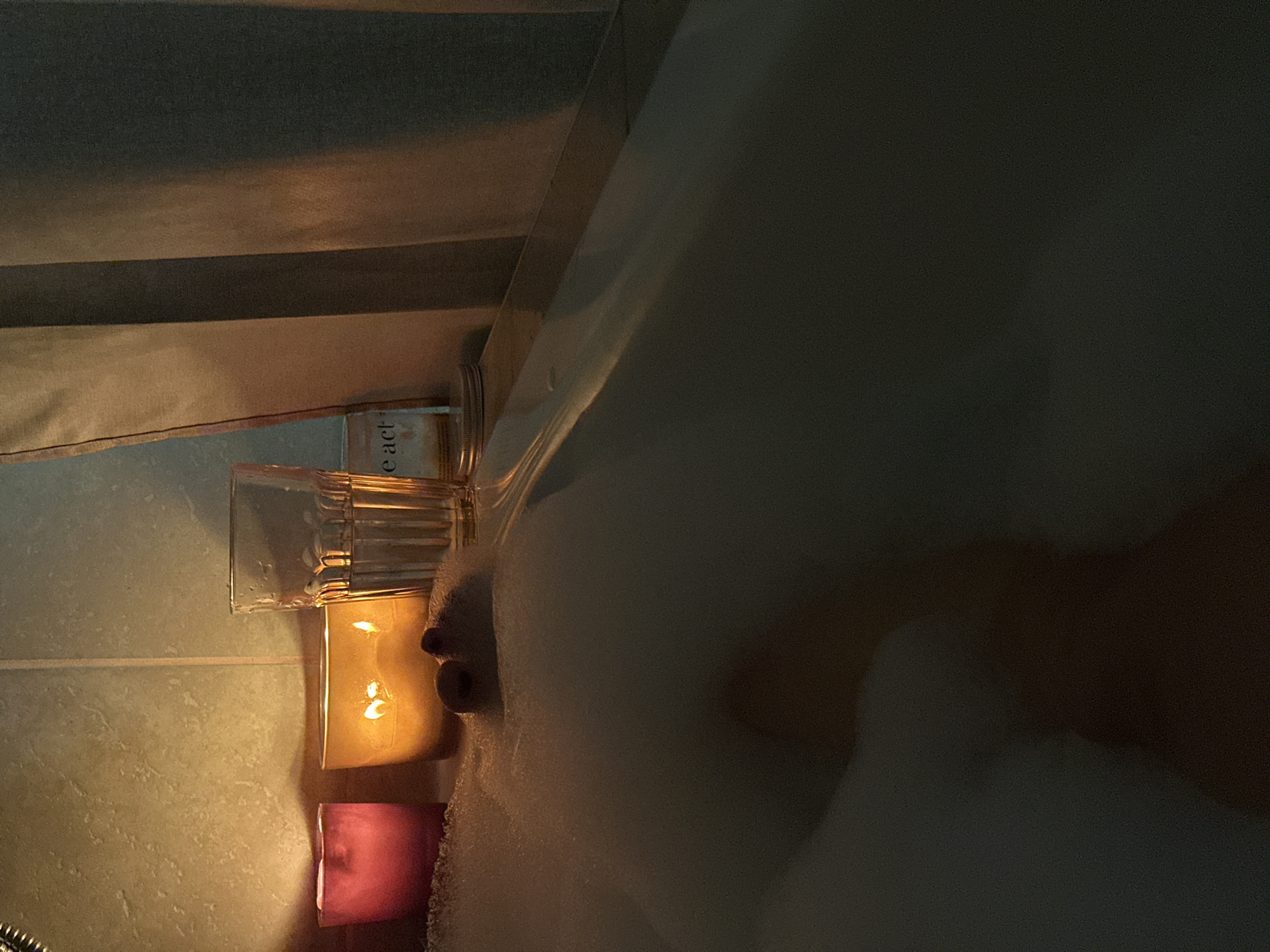 A dimly lit bathroom scene. A bathtub filled with bubbles occupies the right side of the image. Two candles, one yellow and one pink, are lit on the side of the tub next to a glass. There is part of a human face peaking out of the bathtub bubbles. The wall is tile with a horizontal stripe. It's unknown if this image was taken in Chaikovsky, Russia.