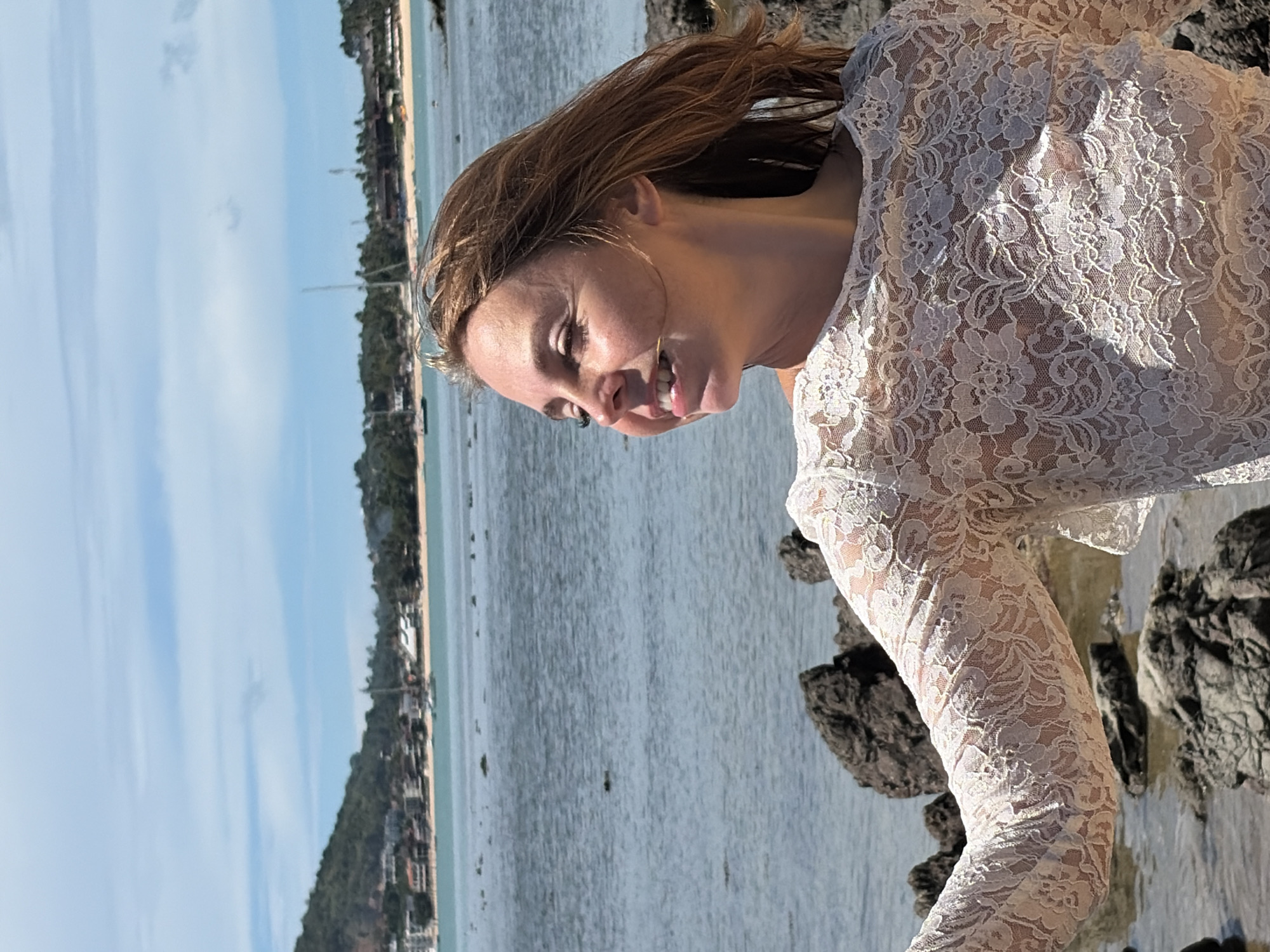 A medium shot shows a Caucasian woman in her late 20s or early 30s from the chest up, standing on a rocky shore with the ocean and a distant coastline in the background. The woman is facing to her right, with her head turned slightly towards the camera, and is smiling. Her eyes are closed, and her hair, which is light brown and windswept, frames her face. She is wearing a white, lace, see-through blouse. The background shows a calm, blue body of water, with a shoreline featuring buildings and lush green hills. Several sailboats are visible on the water. The scene appears to be in a tropical location, possibly Baan Bo Phut, Thailand, given the coastal environment and vegetation. The lighting suggests it is daytime, with bright sunlight casting shadows. The rocks in the foreground are dark and uneven.