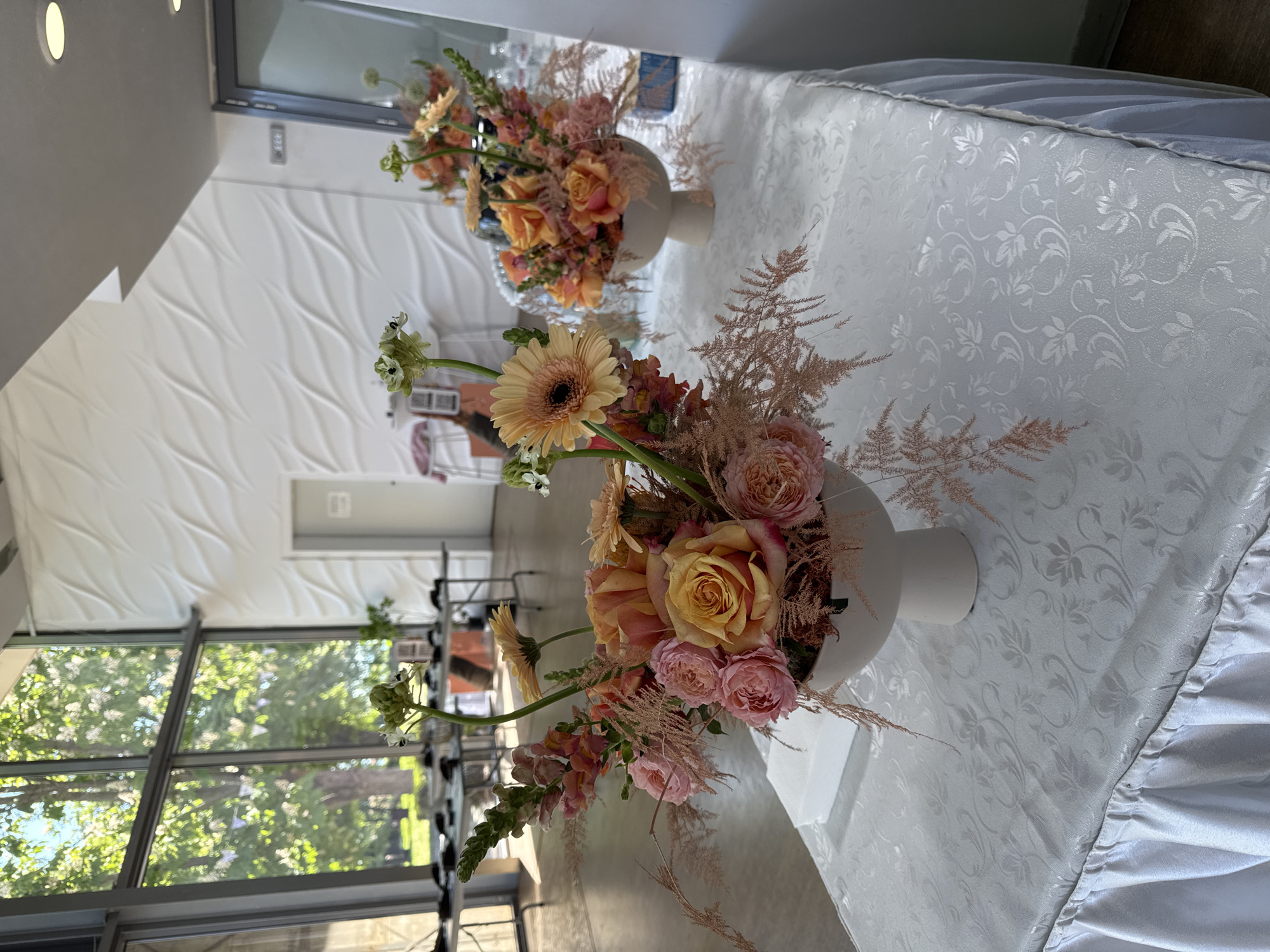 The image shows two floral arrangements in white, round vases on a white tablecloth with a floral pattern and a ruffled edge. The arrangements consist of flowers in shades of orange, pink, and green, including roses, gerberas, and other flowers. The background features a window with a view of trees and sky, suggesting an outdoor setting. The interior includes a white wall with a textured, wavy pattern. Based on the details, the location seems to be an event venue with large windows and decorative walls, likely in Burgas, Bulgaria.