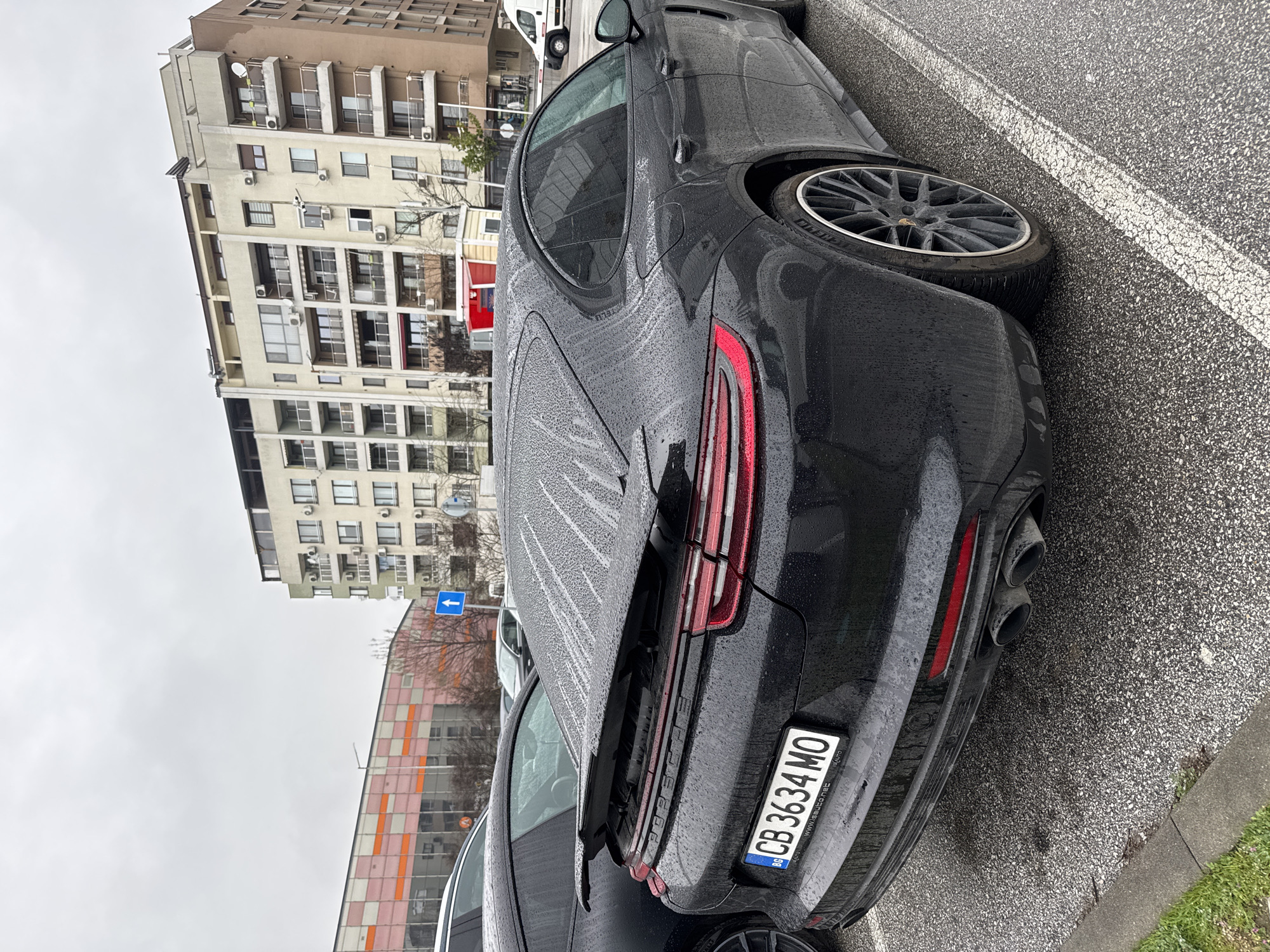 The image captures the rear view of a black Porsche Panamera parked on a city street. The car is wet, indicating recent rainfall. The license plate reads "CB 3634 MO," indicating registration in Bulgaria. The car is parked on an asphalt surface marked with white parking lines. In the background, several multi-story buildings are visible, characteristic of an urban environment. The presence of air conditioning units on the building facades suggests residential or commercial use. Based on the architectural style and the license plate, the location is likely Plovdiv, Bulgaria. The weather appears overcast. There are no people visible in the immediate vicinity of the car.