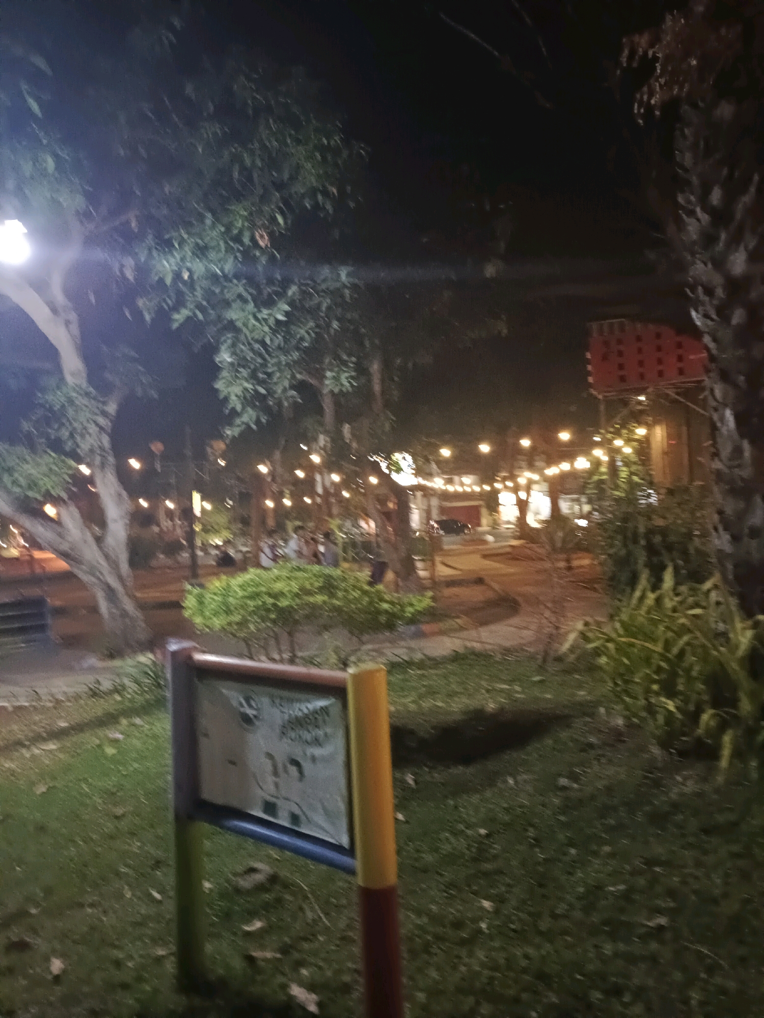 The image depicts a nighttime scene in what appears to be a park or public gathering space in Lamongan, Indonesia, likely near a food court or restaurant area. String lights are visible, illuminating trees and pathways. A signpost with a notice board stands in the foreground, its text partially obscured and unreadable. The environment is characterized by lush greenery, including trees and bushes, with a well-maintained grassy area. The lighting suggests a lively evening atmosphere, possibly a recreational or dining destination for locals and visitors.