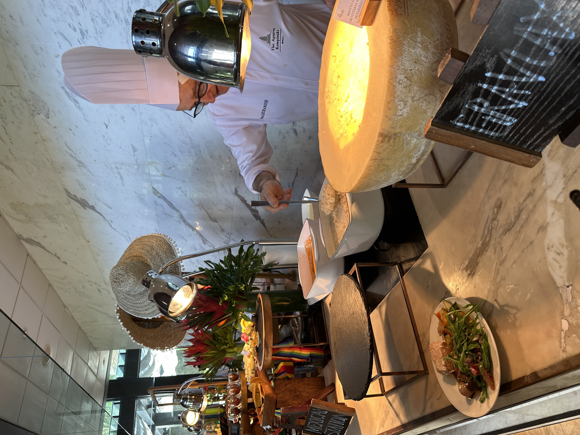 The image depicts a chef in a white uniform and chef's hat, preparing risotto in a large Parmesan cheese wheel at The Apurva Kempinski Bali in Nusa Dua, Indonesia. The cheese wheel is lit by a heat lamp above. The scene is set on a marble counter, with a chalkboard menu displayed nearby. Other food items, including a plate of salad and a "Mexican Station", are visible in the background.