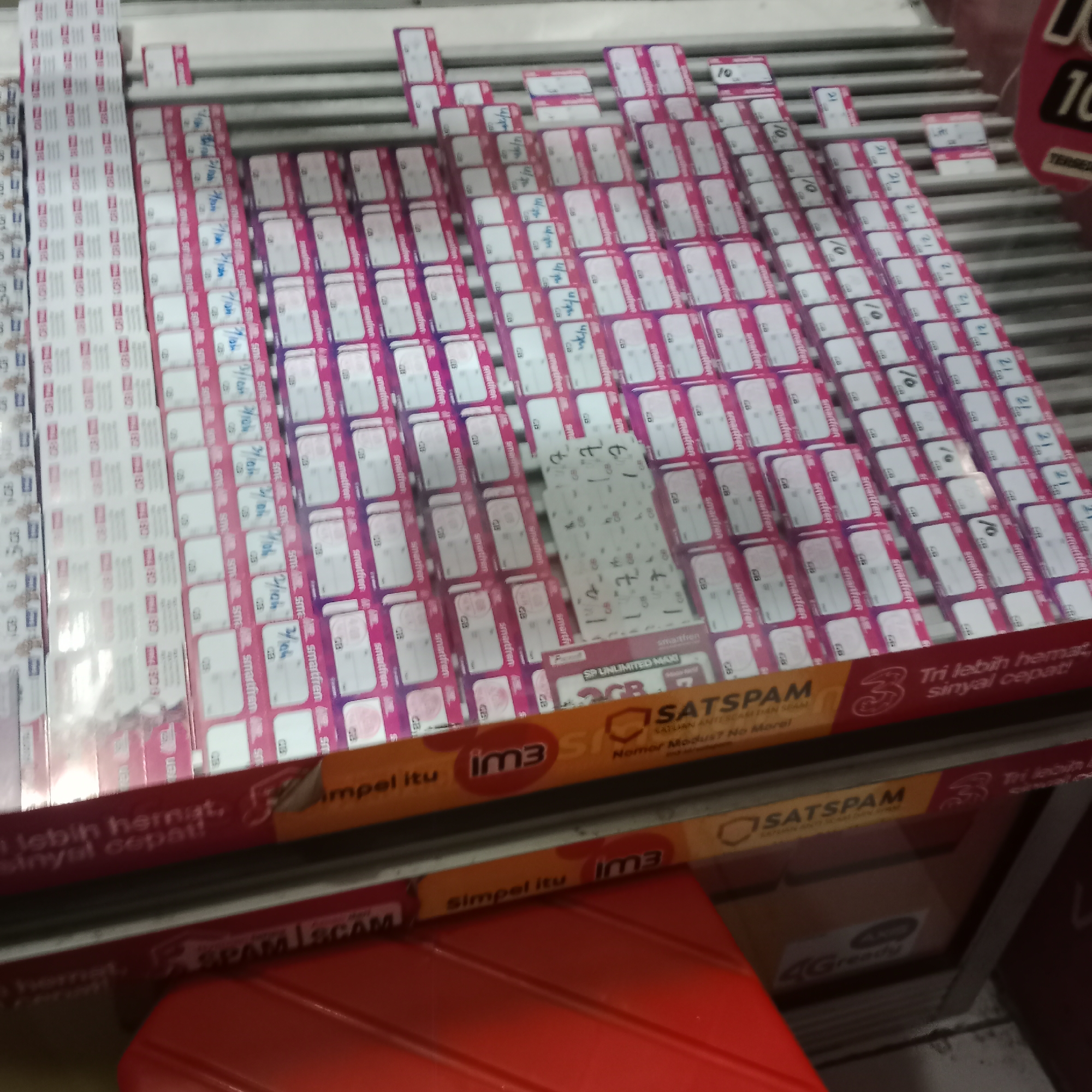 The image shows a display of mobile phone SIM cards in what appears to be a retail setting in Tegowanu Kulon, Indonesia. The SIM cards are neatly arranged in rows on shelves. Many of the SIM card packages are pink and white, with some having purple accents. Text and logos are visible on the packaging, including "smartfren," "10," "21," and "3 GB." Some of the packages also have handwritten notations. In the foreground, there are promotional banners with text like "Tri lebih hemat, sinyal cepat!" and "SATSPAM." The overall impression is that of a mobile service provider's point of sale.