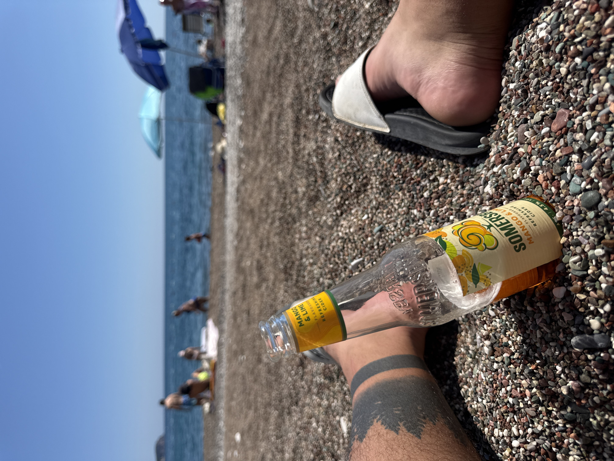 The image captures a first-person perspective on a pebbly beach in Rafailovići, Montenegro, under a clear, bright sky. In the immediate foreground, a person's legs and feet are visible, resting on the multi-colored pebble surface. The left leg displays a prominent black band tattoo, suggestive of a forest or mountain landscape at its base. A partially consumed Somersby Mango & Lime flavored refreshing cider bottle, with its yellow and green label clearly legible, rests upright on the pebbles between the person's legs. The right foot wears a white-strapped, black slide sandal.
The midground, intentionally blurred, reveals several individuals on the beach near the sea. Visible figures include adults standing and seated, and at least one child. Some are near the water's edge, while others appear to be relaxing on towels or mats. Two beach umbrellas are discernible: one light blue and one dark blue. Beyond them, the Mediterranean Sea extends under a clear, light blue sky. A distant landmass or island is faintly visible on the horizon to the left. The overall scene depicts a sunny day at a populated beach.
