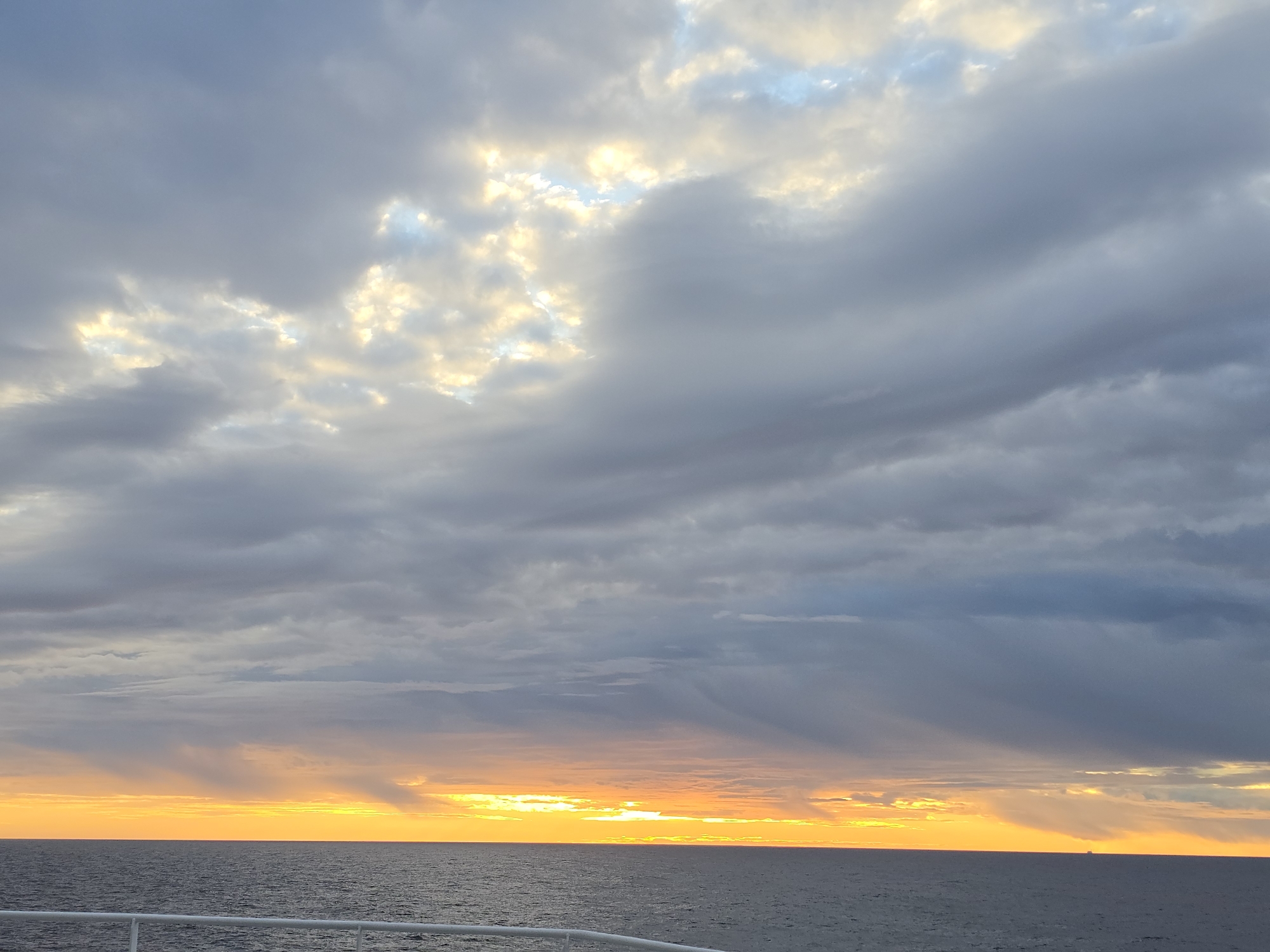 The image captures a sunset over the ocean. The sky is dominated by a thick layer of gray clouds, with some areas where the sun is breaking through, creating a golden glow. The horizon line is visible where the sky meets the dark blue ocean. A small white railing is visible in the bottom left corner of the image. The location in the city of Randaberg, Norway, is not directly discernible from the image but presumed based on context. No people, subjects, actions, or interactions are visible. The environment consists of the sky and the ocean, with the light of the setting sun creating a contrast with the dark clouds. A barely visible ship is positioned on the horizon.