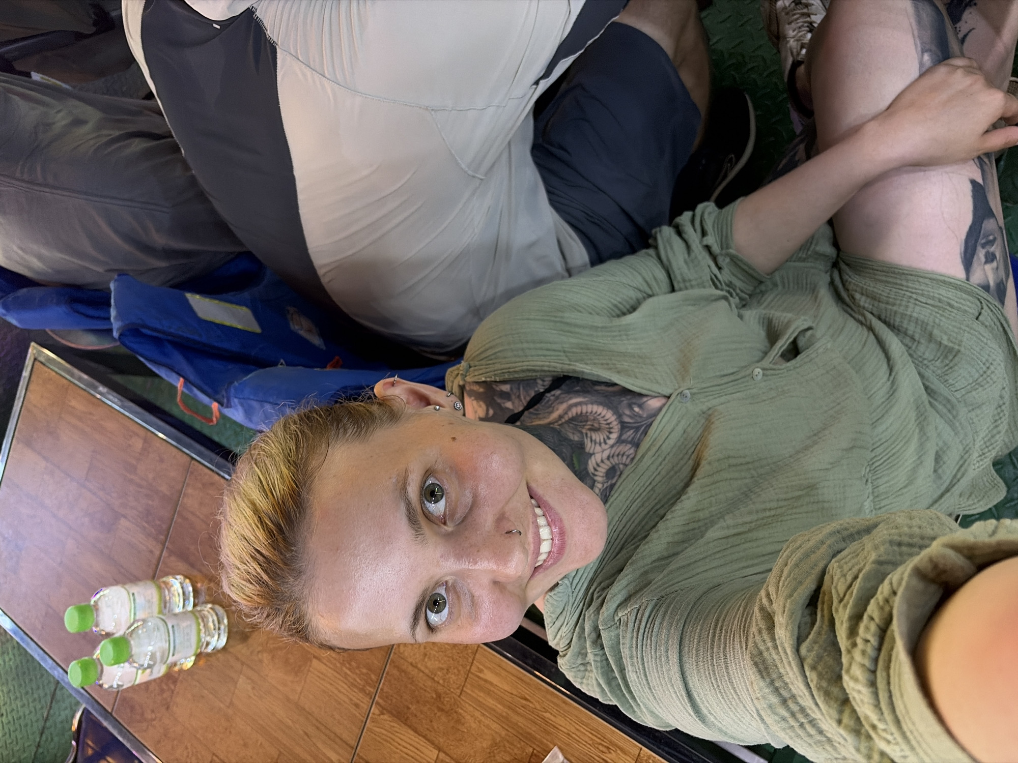 This media file captures a close-up, slightly tilted selfie of a smiling woman in a casual setting, likely in Đà Nẵng, Vietnam.
**Subjects:** The primary subject is a woman with light-colored hair, green eyes, a nose ring, and multiple ear piercings. She has extensive tattoos visible on her neck, upper chest, and left thigh. She is wearing a loose-fitting, light green, textured long-sleeved shirt. Parts of another person are visible in the background, showing their legs and torso, dressed in grey, white, and dark blue athletic-style clothing (shorts, top, and sneakers).
**Objects:** In the foreground, four plastic water bottles with green caps are resting on a wooden table. A blue fabric item, possibly a bag or life vest, is visible next to the table. The floor appears to be a dark green, possibly patterned, surface.
**Scene and Setting:** The scene takes place in what appears to be an indoor or covered area, given the consistent lighting and the presence of a table. The wooden table and the casual attire of the subjects suggest a relaxed environment, potentially a café, restaurant, or a mode of transport like a boat.
**Activity or Event:** The woman is actively taking a selfie, looking directly at the camera with a bright, relaxed smile. The other person seems to be seated and resting.
**Notable Details:** The woman's expression conveys happiness and contentment. Her visible tattoos are a prominent feature. The water bottles suggest a break or travel activity. The location context of Đà Nẵng, Vietnam, aligns with the casual dining or travel atmosphere suggested by the elements in the image. No specific time of day is explicitly indicated, but the lighting suggests daytime.
**Visible Text:** Small, unreadable text can be seen on the labels of the water bottles and on the blue fabric item in the background.