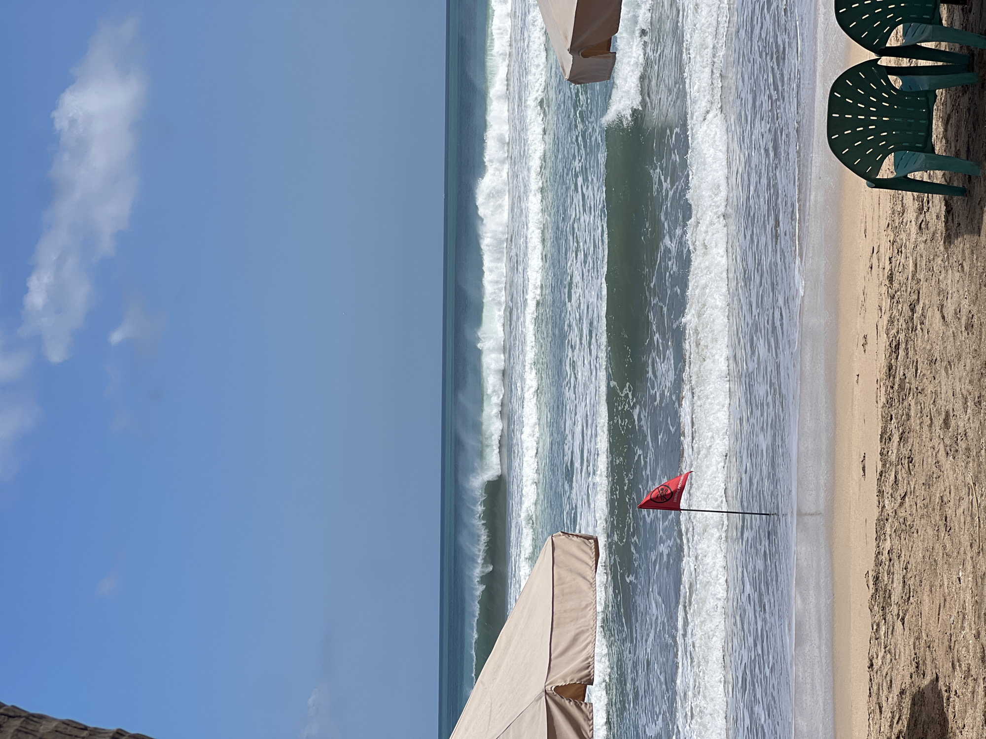 In Kuta, Indonesia, a beach scene unfolds under a bright blue sky dotted with white clouds. Large waves crash onto the sandy shore, creating a dynamic seascape. Two tan beach umbrellas stand on the sand, providing shade. In the foreground, two green plastic chairs are positioned facing the ocean. A red flag, planted in the shallow water, bears the words "SWIMMING PROHIBITED" in white lettering. The overall impression is one of a lively beach with conditions that may not be safe for swimming.
