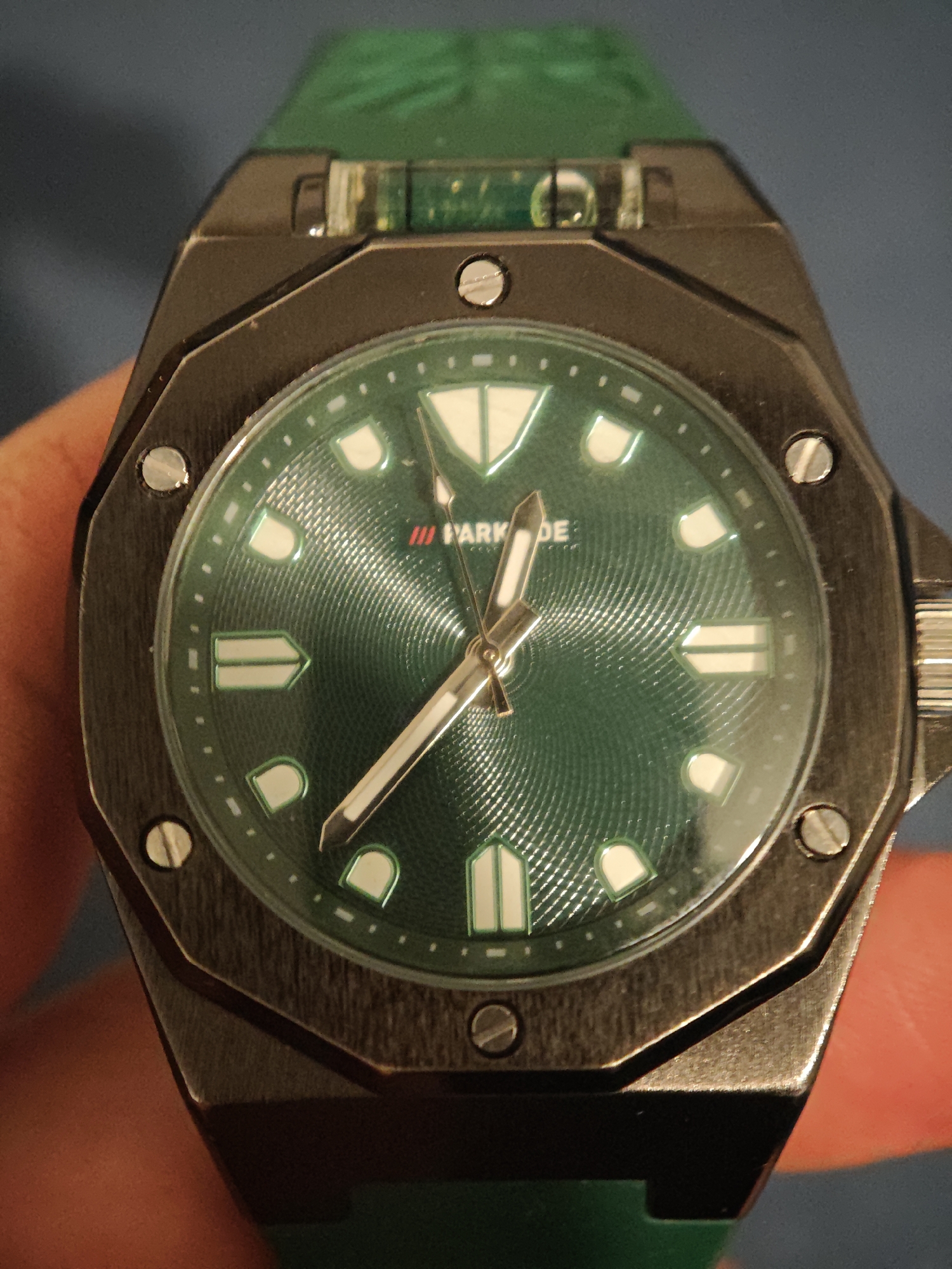 A close-up shot depicts a men's wristwatch. The watch has a dark, possibly gunmetal grey, octagonal bezel secured with visible screws. The watch face is green with a textured, spiral-like pattern. The hour markers are white with a luminous appearance. The watch hands are silver-colored. The watch has a green strap. The text on the watch face reads "III PARK DE" The background is a muted blue.
There is no information available within the image indicating the location is Sofia, Bulgaria.