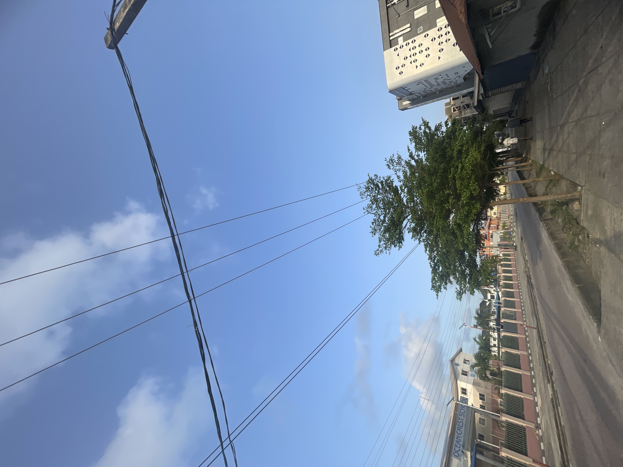 Ilado, Nigeria. A street view shows a road running parallel to a fence. Buildings stand on either side of the road. Overhead, power lines crisscross the clear, blue sky with scattered clouds. A tall, modern building with a white facade and patterned openings stands on the left. A tree with dense green foliage sits along the sidewalk. A building with "OCEAN CREST SCHOOL" sign is visible. People are visible on the sidewalk.