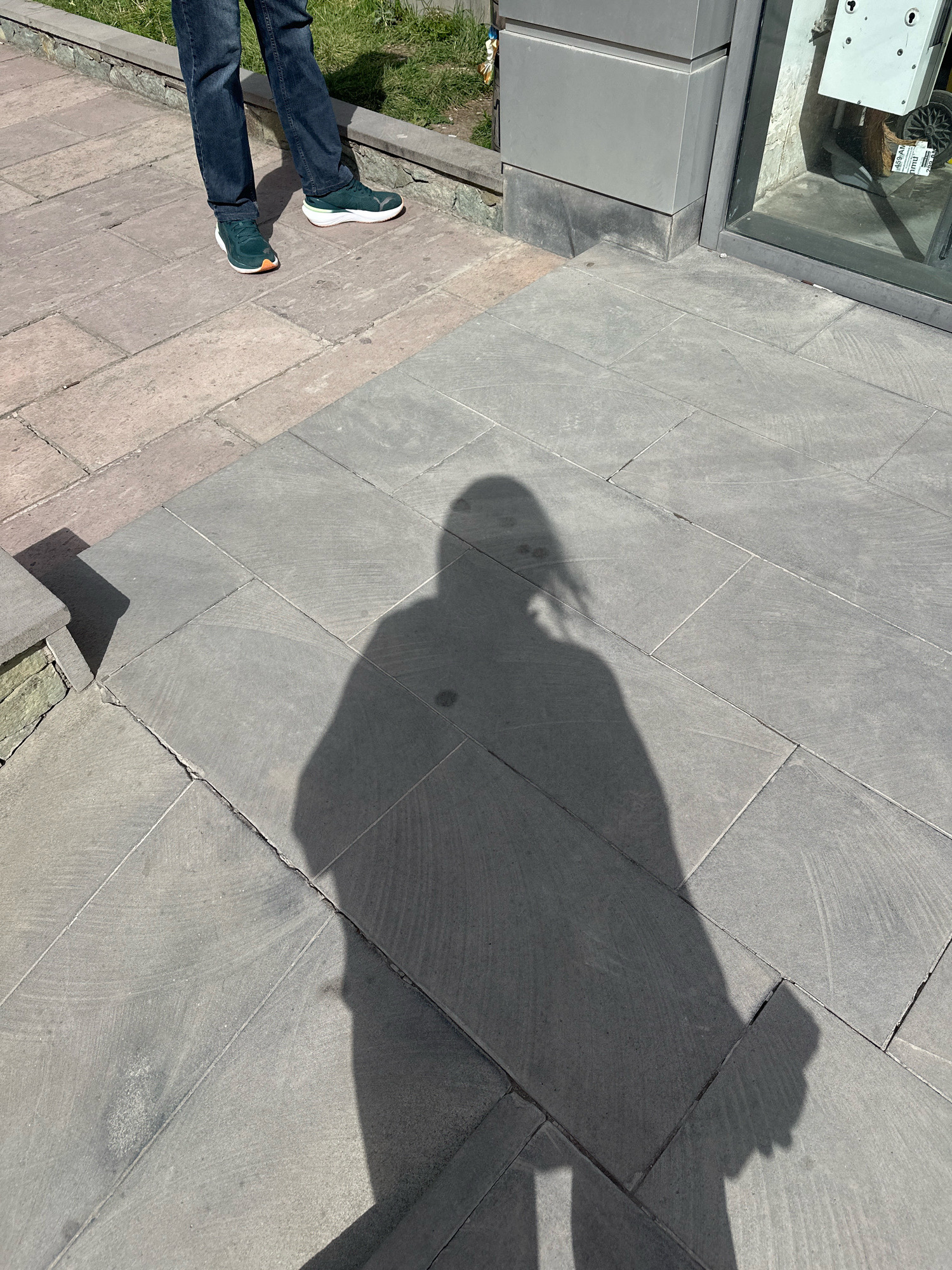 The image shows the shadow of a person cast on a paved walkway. The walkway is made of large, grey stone tiles. To the left, a person's legs are visible, wearing dark jeans and green and orange athletic shoes. They are standing on a different section of paving, possibly an adjacent path or a step, which is made of reddish-brown tiles. The background shows a grassy area and a modern building facade with a glass window. The scene appears to be outdoors, likely in an urban environment. The shadow suggests the sun is positioned to the left and slightly behind the photographer. The overall impression is a casual moment captured in daylight, with a focus on the interplay of light and shadow on the textured surfaces of the paving. This scene could be located in Gyumri, Armenia, or any city with similar paving and architectural styles.