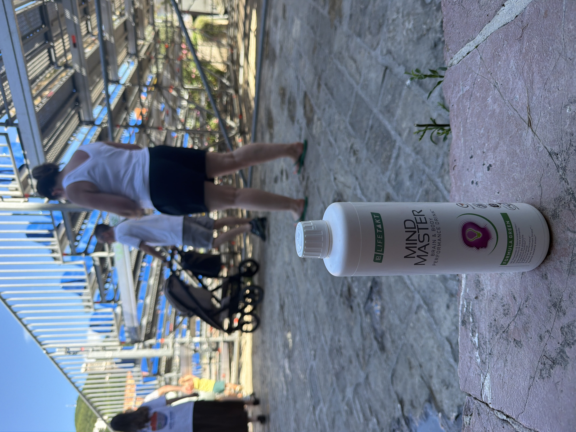 In Budva, Montenegro, a white bottle of "Mind Master Brain & Body Performance Drink" with a green label is placed on a stone-tiled surface in the foreground. The bottle is positioned to the right, partially obscuring the details of the background. In the blurred background, several people are visible, some walking and others standing near a structure that appears to be temporary seating or scaffolding, possibly at an outdoor event or stadium. A stroller is also present. The sky is clear and blue, indicating a sunny day. The overall scene suggests an outdoor public space in Budva.