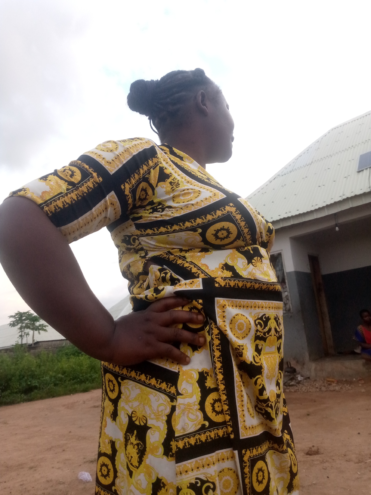 The image depicts a woman standing outdoors in Serti, Nigeria. She is positioned in the foreground, facing right and looking away from the camera. The woman is wearing a black and gold patterned dress, and her right hand is resting on her hip. In the background, a building with a corrugated metal roof and a solar panel is visible. Further in the distance, there are trees and vegetation. The ground is a dirt path. The sky is overcast.