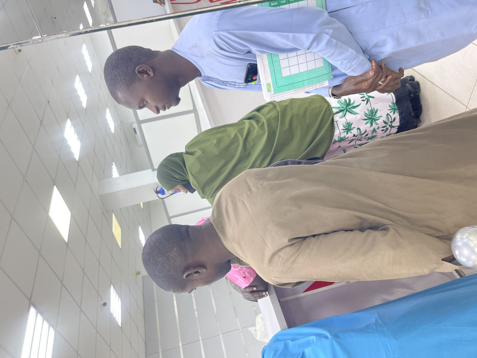 The image depicts an indoor public service facility, possibly in Zaria, Nigeria, characterized by white walls, a white ceiling with square panels, and evenly spaced fluorescent lighting. A black security camera is visible on the ceiling in the upper left.
Three individuals are primarily visible, along with a partial view of a fourth. From left to right:
1. A person in a light blue garment is partially visible, facing right, near a grey-topped counter.
2. A male individual stands with his back to the camera, wearing a light brown, long-sleeved tunic. He leans slightly over the grey counter, with his right arm bent at the elbow and resting on its surface.
3. A female individual stands behind the counter, positioned with her back mostly to the camera, but slightly turned towards the right. She wears a dark olive green hijab covering her head and shoulders, and a white skirt with a repeating green leaf pattern. A pink object is held in her visible right hand.
4. A male individual stands in profile on the right, facing right. He has short dark hair and wears a light blue traditional tunic with embroidered details on the chest. He holds a light green-bordered notebook or folder tucked under his left arm. A dark-colored smartphone is partially visible in a pocket on his right side. His hands are clasped in front of him.
All individuals are standing. The environment suggests a waiting area or a service point. A reflective surface, possibly a mirror or glass partition, is present on the far right edge of the frame. A silvery, spherical object is visible at the bottom center of the image.