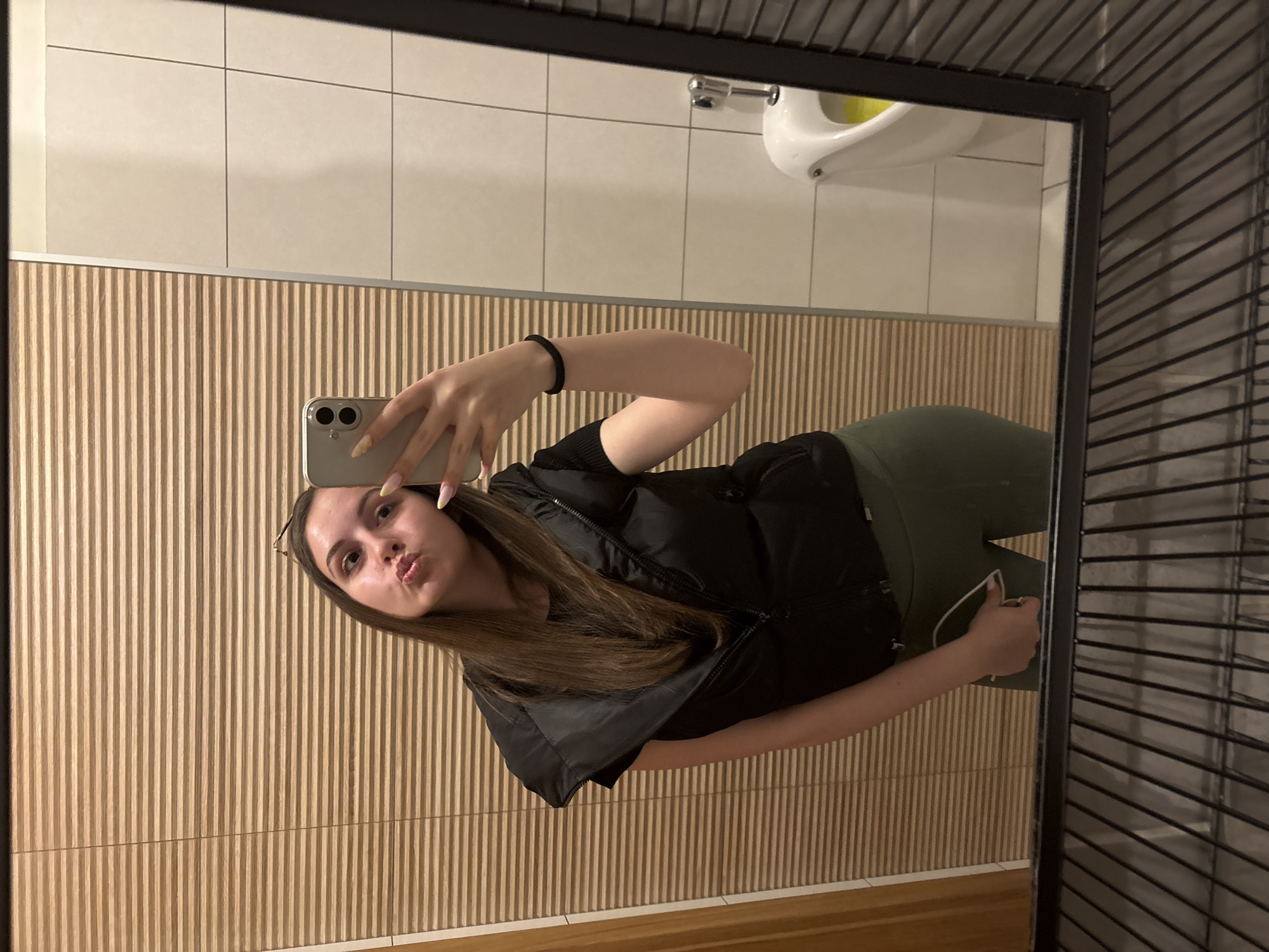 A woman is taking a selfie in a mirror located in a bathroom. The bathroom has beige tile walls with vertical ridges, and a white sink is visible in the background. The woman is wearing a black vest and olive green leggings. The location is presumed to be in Burgas, Bulgaria, although there is no specific indication to support this.