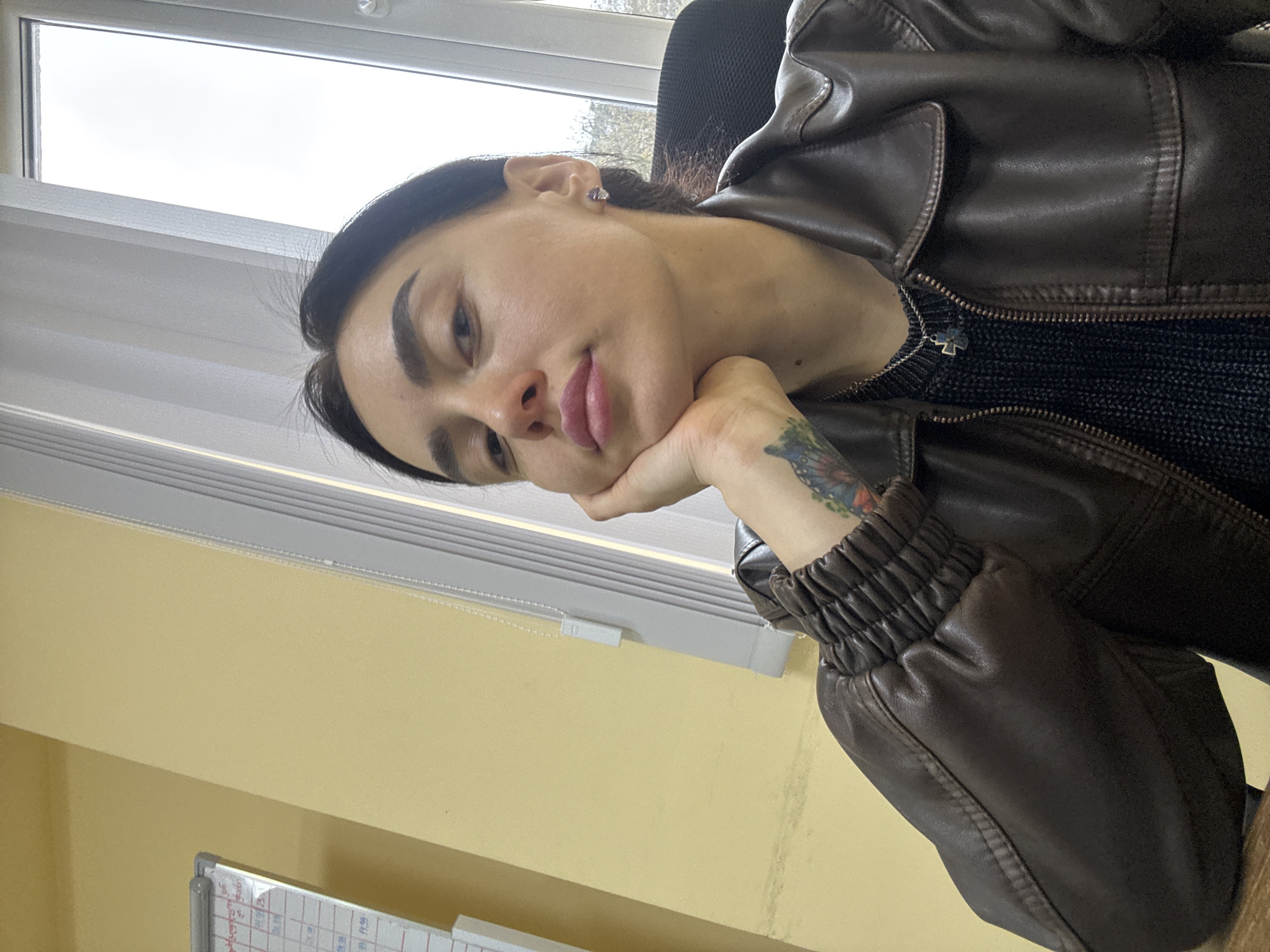 A woman is indoors, resting her head on her hand. She has dark hair and eyebrows, wearing a brown leather jacket over a black sweater. A small silver cross hangs from her neck. A tattoo is visible on her forearm. The background shows a window with blinds and a yellow wall. A whiteboard with writing is partially visible in the lower left corner. The location appears to be indoors, possibly an office in Tbilisi, Georgia, given the setting and subject's possible location.