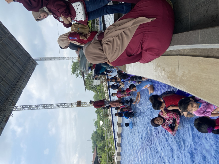 A group of children play in a blue-tiled swimming pool. Some are standing, others are wading, and some are submerged in the water. Several adults stand at the pool's edge, watching them. The area appears to be an outdoor recreational space with metal support beams visible. The sky is light blue with scattered clouds.