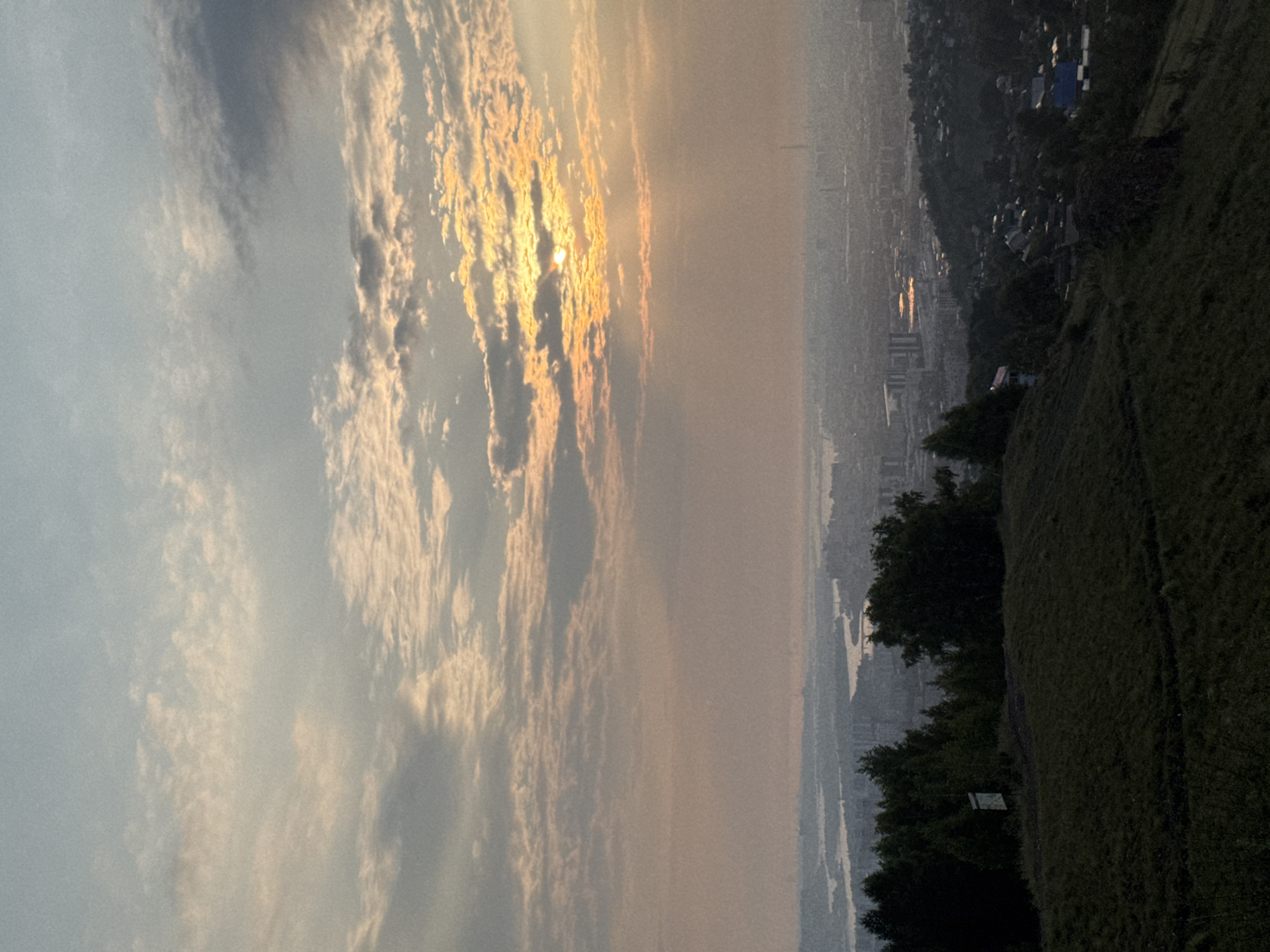 The image captures a panoramic view of Krasnoyarsk, Russia, at sunset, as seen from the Nikolskaya Sopka hill. The sky is filled with scattered clouds, tinged with the warm hues of the setting sun, which is partially obscured by the clouds. Rays of sunlight break through, illuminating the city in the distance. The cityscape is densely populated with buildings of varying heights, interspersed with green spaces and a winding river that reflects the sky's colors. In the foreground, the grassy slope of Nikolskaya Sopka is visible, with scattered trees and a signpost. The overall atmosphere is serene, with the setting sun casting a soft, diffused light over the urban landscape.