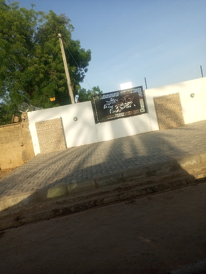 The image shows a white concrete wall with a black metal gate. The gate is decorated with a floral pattern. There are two light fixtures on the wall, one near the gate and another near a door. The door is made of wood with horizontal slats. The wall is built next to a dirt road. There is a concrete sidewalk in front of the wall. The road and sidewalk are covered with shadows. The sidewalk is made of grey bricks arranged in a pattern. There is a tree to the left of the wall, casting a shadow. The scene is taken in Maiduguri, Nigeria, at the coordinates 11.817 latitude and 13.153 longitude.