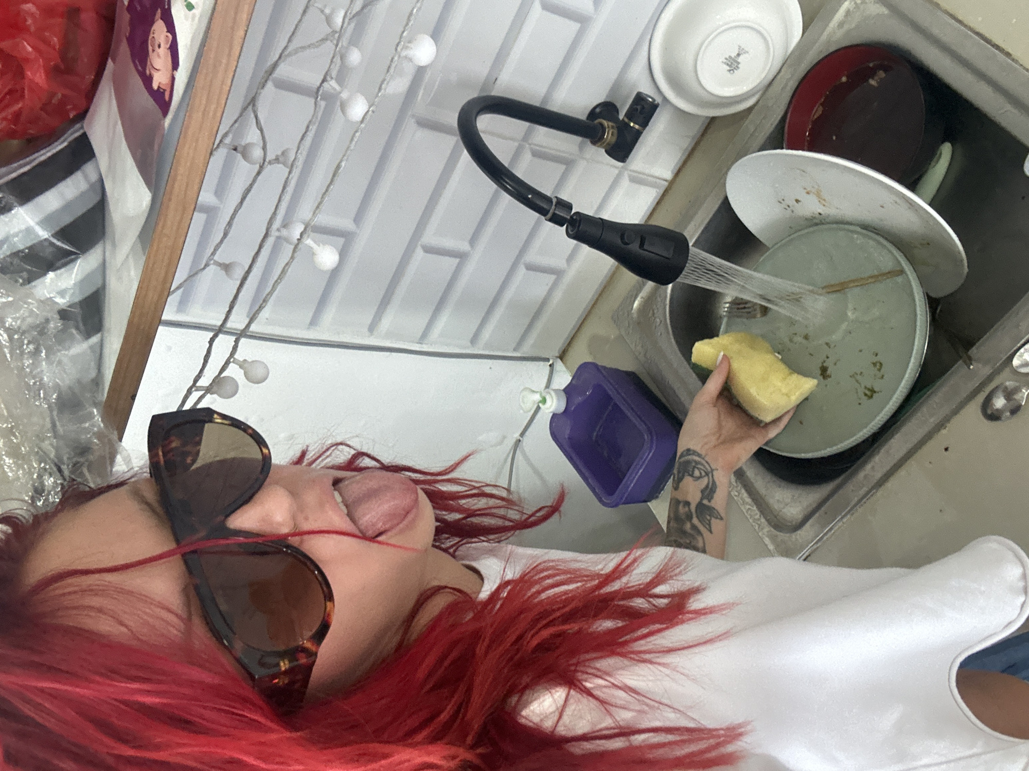 This image captures a person engaged in dishwashing in a kitchen, likely in North Kuta, Indonesia, given the provided context. The perspective is from above, looking down at the person and the sink area.
The main subject is a person with strikingly bright red hair, wearing a white top. They have tortoiseshell-patterned sunglasses pushed up onto their forehead, and their tongue is playfully sticking out, suggesting a lighthearted or silly mood. A distinct black tattoo, depicting an eye and part of a face, is visible on their left forearm as they hold a yellow sponge.
The activity taking place is washing dishes. Water streams from a modern, black flexible kitchen faucet onto a light green plate, which the person is cleaning with the sponge. The metallic sink is filled with several dirty dishes, including a red plate and multiple white plates and bowls. One white bowl at the top left of the sink has an unreadable logo on its base.
The background features a white tiled backsplash. To the left of the sink, there is a purple plastic container or dispenser with a white pump. In the upper left corner of the image, various items are visible, including a red plastic bag, a striped bag, and a wooden frame. White decorative string lights with round bulbs are strung across the top left portion of the scene. No discernible text is visible on any objects in the image.