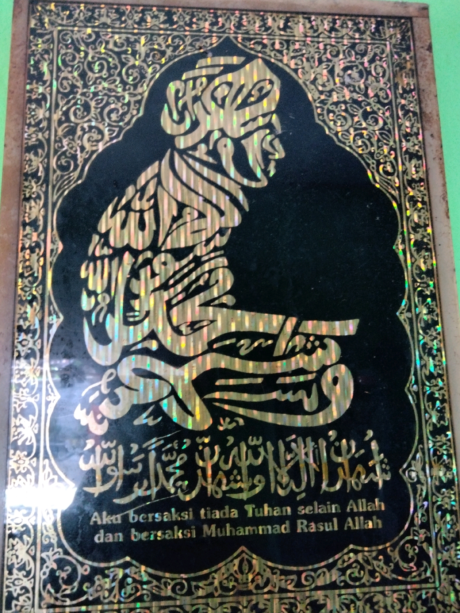 The image displays a close-up view of a book cover with intricate golden Arabic calligraphy on a black background. The calligraphy depicts a kneeling figure, likely a religious or spiritual representation. Below the Arabic script, there is text in Indonesian: "Aku bersaksi tiada Tuhan selain Allah dan bersaksi Muhammad Rasul Allah". This translates to "I bear witness that there is no god but Allah, and I bear witness that Muhammad is the Messenger of Allah." The book cover is adorned with ornate borders featuring floral and geometric patterns, also in gold foil, which exhibit holographic qualities, reflecting iridescent colors. The overall impression is one of religious significance and artistic embellishment. The image itself does not contain any elements that would allow for the determination of its specific location within Rahong, Indonesia, as it is a close-up of an object with no discernible environmental context beyond the object itself and a hint of a greenish background.
