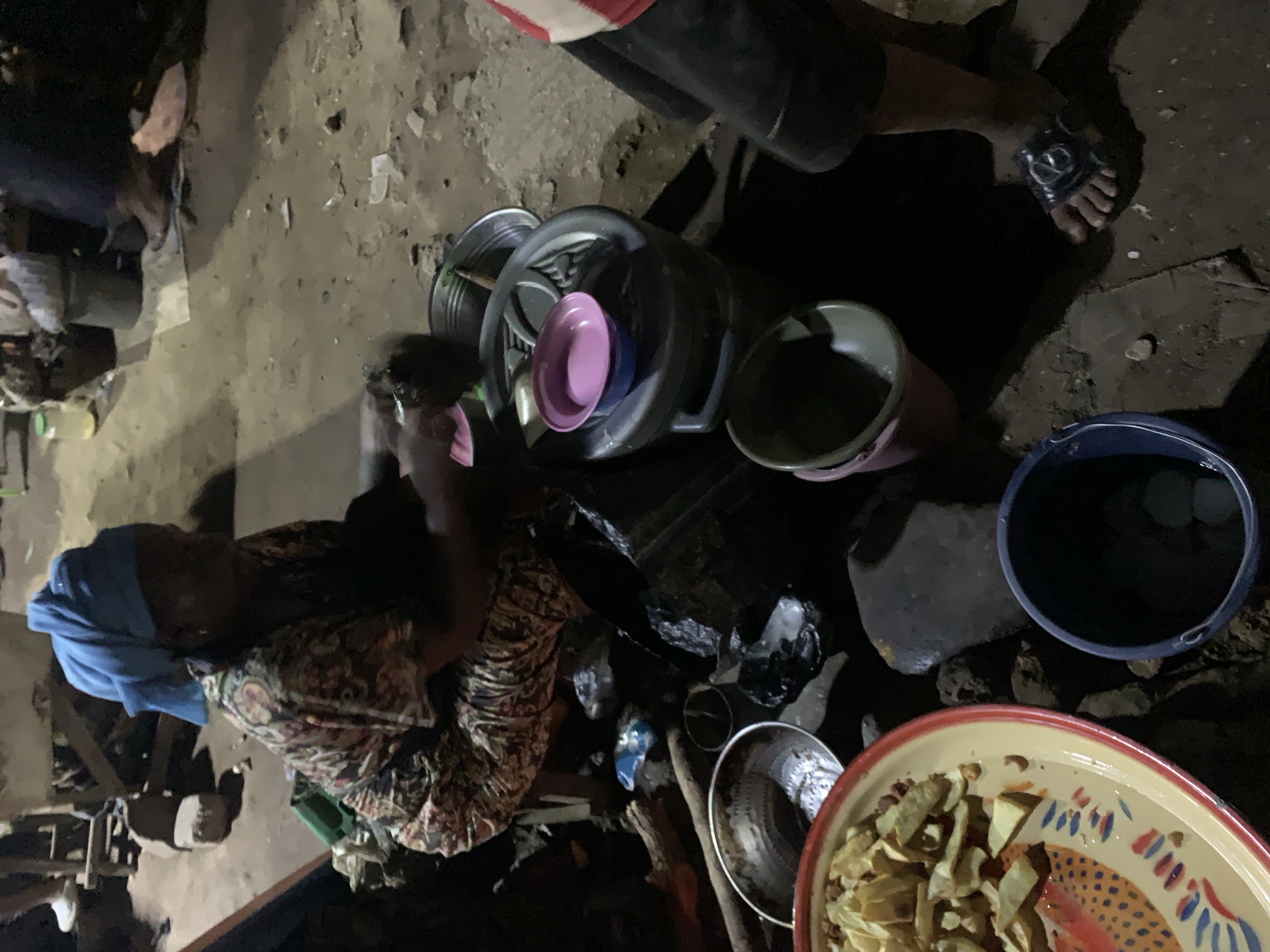 In Rafin Par, Nigeria, a person wearing a blue headscarf and patterned dress is seated near a collection of kitchenware, including metal pots, plastic buckets, and a large decorative bowl filled with food. Another person's lower torso and feet in sandals are visible. The scene is dimly lit and appears to be outdoors on a rough, uneven surface.