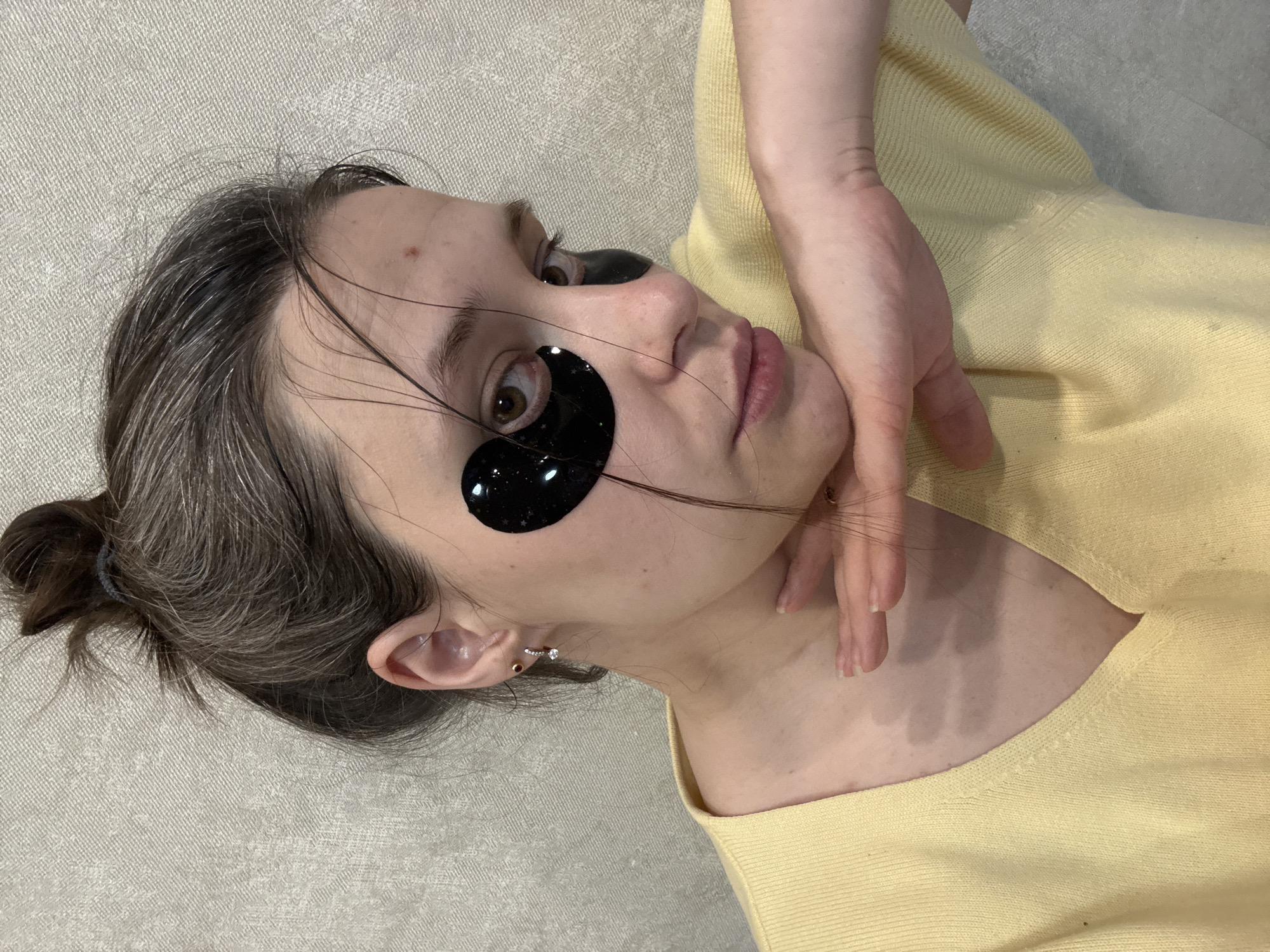 A woman is lying on her side on a light-colored textured surface. She is wearing black eye patches and a light yellow shirt. Her brown hair is pulled back in a bun. Her hand is near her face.
The location in the city of Chaikovsky, Russia is not discernible from the image.
