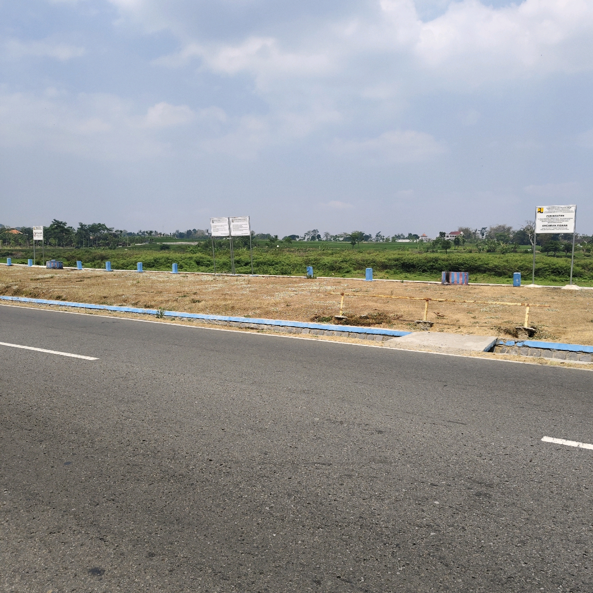 The image shows a roadside view in Botok, Indonesia. A paved asphalt road occupies the foreground, with a white line marking its edge. Adjacent to the road is a dry, grassy area bordered by a low blue-and-white striped concrete barrier. Several blue bollards are spaced along this barrier, and some yellow posts with horizontal bars are also present. In the background, a lush green field stretches out, dotted with trees and some buildings, suggesting a semi-rural or suburban environment. Two white signs are visible on poles in the field, one on the left and another on the right. The sky above is partly cloudy with a clear horizon. The overall scene depicts an outdoor environment with evidence of development or land management.