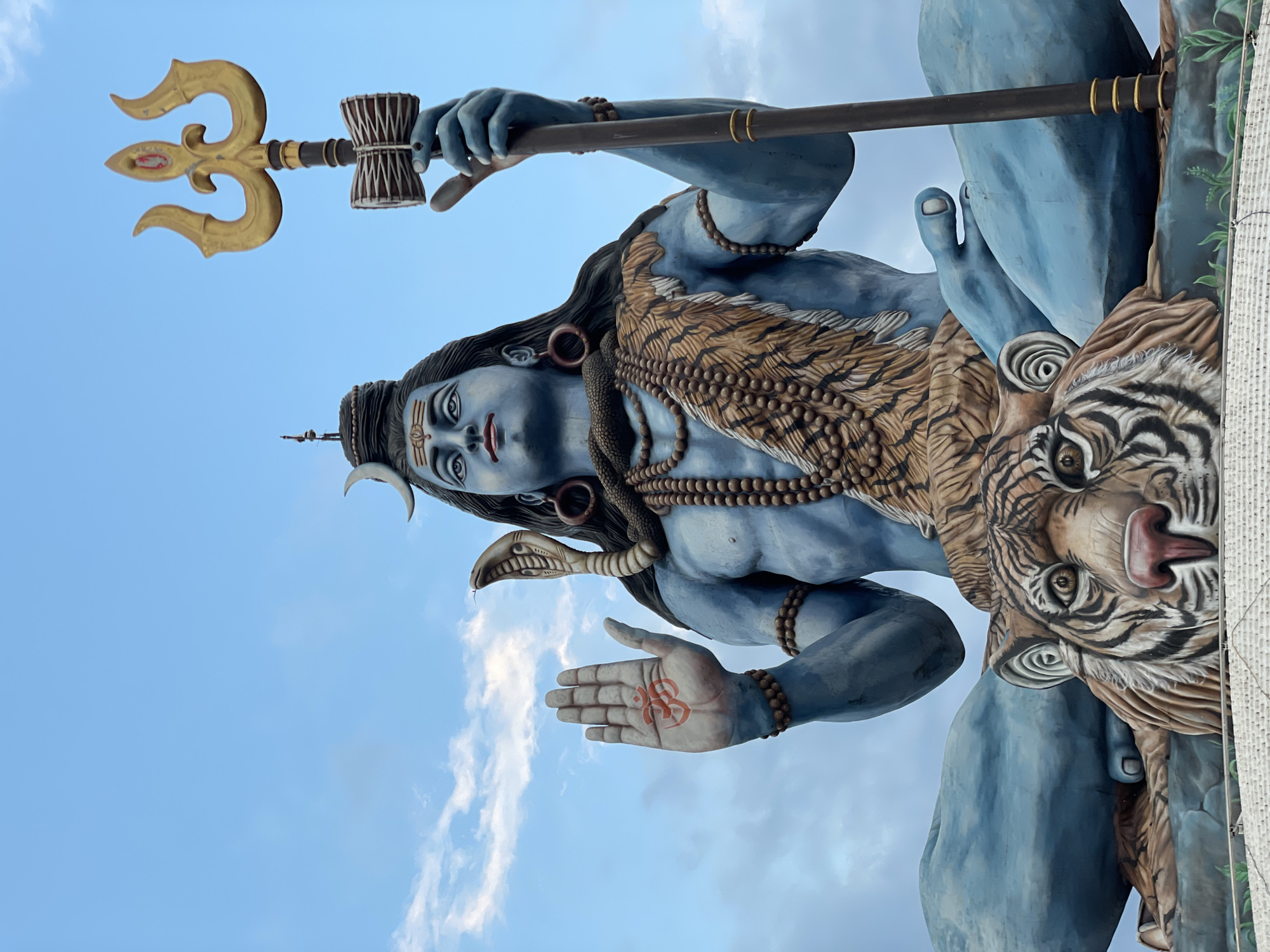 The image depicts a large, blue-skinned statue of the Hindu deity Shiva, prominently featuring a trident, a hand adorned with a symbol, and a tiger. The statue is set against a backdrop of a bright blue sky interspersed with scattered white clouds. The location is identified as Pokhara, Nepal.
