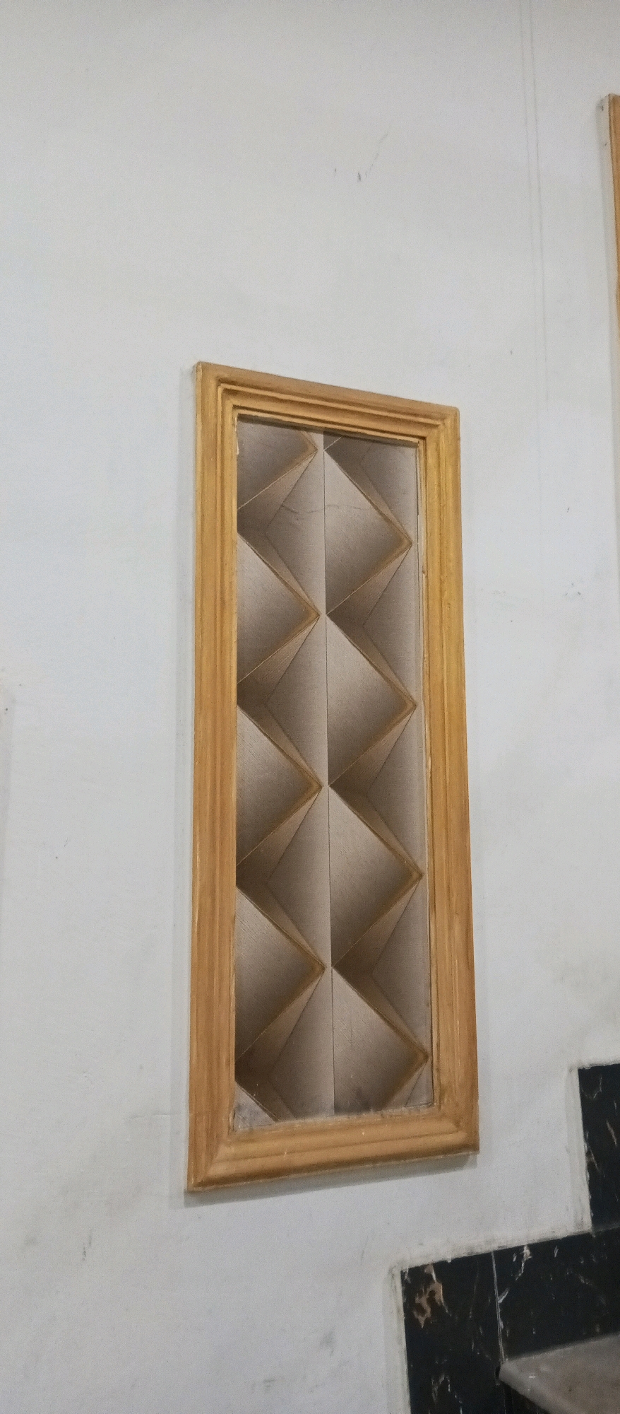A narrow, rectangular window with a wooden frame is set into a white wall. The window is filled with a decorative insert featuring a pattern of interlocking diamond shapes. The insert appears to be made of a light-colored material with a textured or folded surface, creating a sense of depth and shadow. The wall surrounding the window is plain and white, with some faint marks or smudges visible. In the lower right corner of the image, a portion of black and white checkered tile work is visible, suggesting this might be an interior or covered outdoor space. The overall scene is static and architectural. The location provided, Gujranwala City Tehsil, Pakistan, is not visually discernible from the image itself.