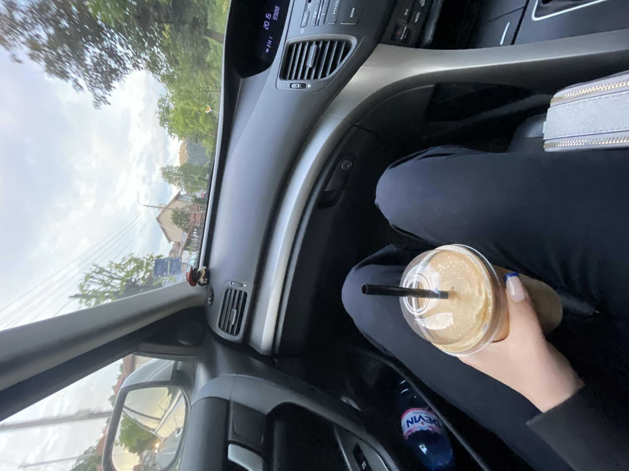 The image depicts the interior of a car, likely in motion. A hand holds a plastic cup with a straw, containing a beverage that appears to be a coffee drink. A bottle of Evian water is visible near the drink. The person is wearing black clothing. The car's dashboard and interior features are partially visible, including the radio display showing "20 15 FM I," dashboard vents, and an airbag panel. In the background, seen through the windshield and side window, is a blurry urban landscape. Based on the general environment and the presence of what appears to be a small mascot or decoration on the dashboard, the location is potentially Zlatograd, Bulgaria. Buildings, trees, and power lines are discernible in the background.