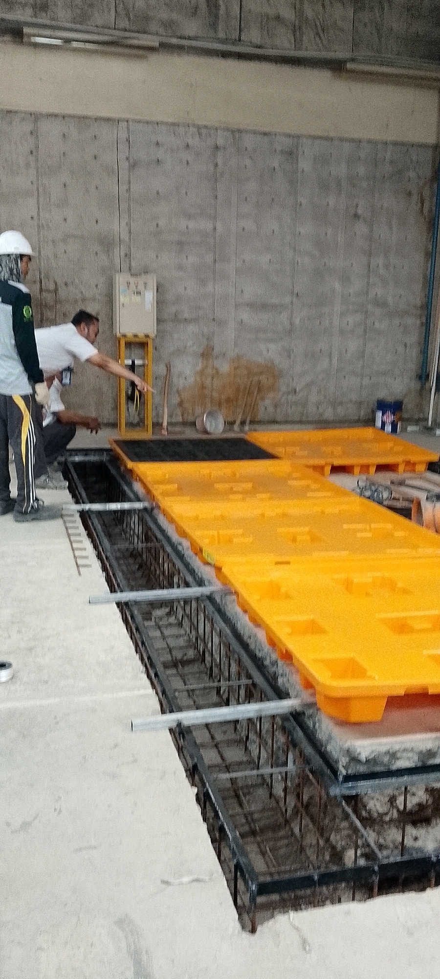 In an indoor construction setting, two individuals are observed near a trench filled with rebar. One person, wearing a white hard hat and a grey and green work uniform, stands on the left. The second person, wearing a white shirt, is kneeling and pointing into the trench. Large, bright orange modular plastic forms, typically used for concrete pouring, are laid out on the ground adjacent to the trench. In the background, a concrete wall with a textured surface dominates the scene. A beige electrical control panel on a yellow stand is positioned against the wall, and various tools, including a shovel and a metal basin, are scattered nearby. A blue bucket with a red and white label is also visible. The floor appears to be concrete.