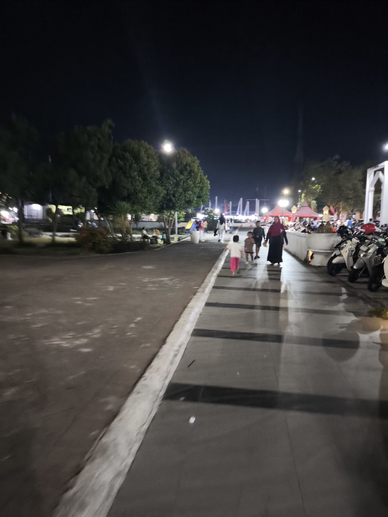 The image depicts a public area at night in Lumajang, Indonesia, likely a park or plaza. In the foreground, a paved walkway with a concrete curb is visible. To the left, a wide, unpaved area with scattered debris and sparse vegetation extends towards trees and some illuminated structures in the background.
On the paved walkway, a group of people are walking. A woman in a red hijab and dark clothing is in the lead, followed by two children, one wearing a pink shirt and pink pants, and another child behind them. Further back, several other individuals are seen walking, and a line of parked motorcycles is visible to the right of the walkway. The background shows a dimly lit scene with trees, streetlights, and some buildings or market stalls. The sky is dark and clear, indicating it is nighttime. The overall impression is a lively evening scene in a public space.