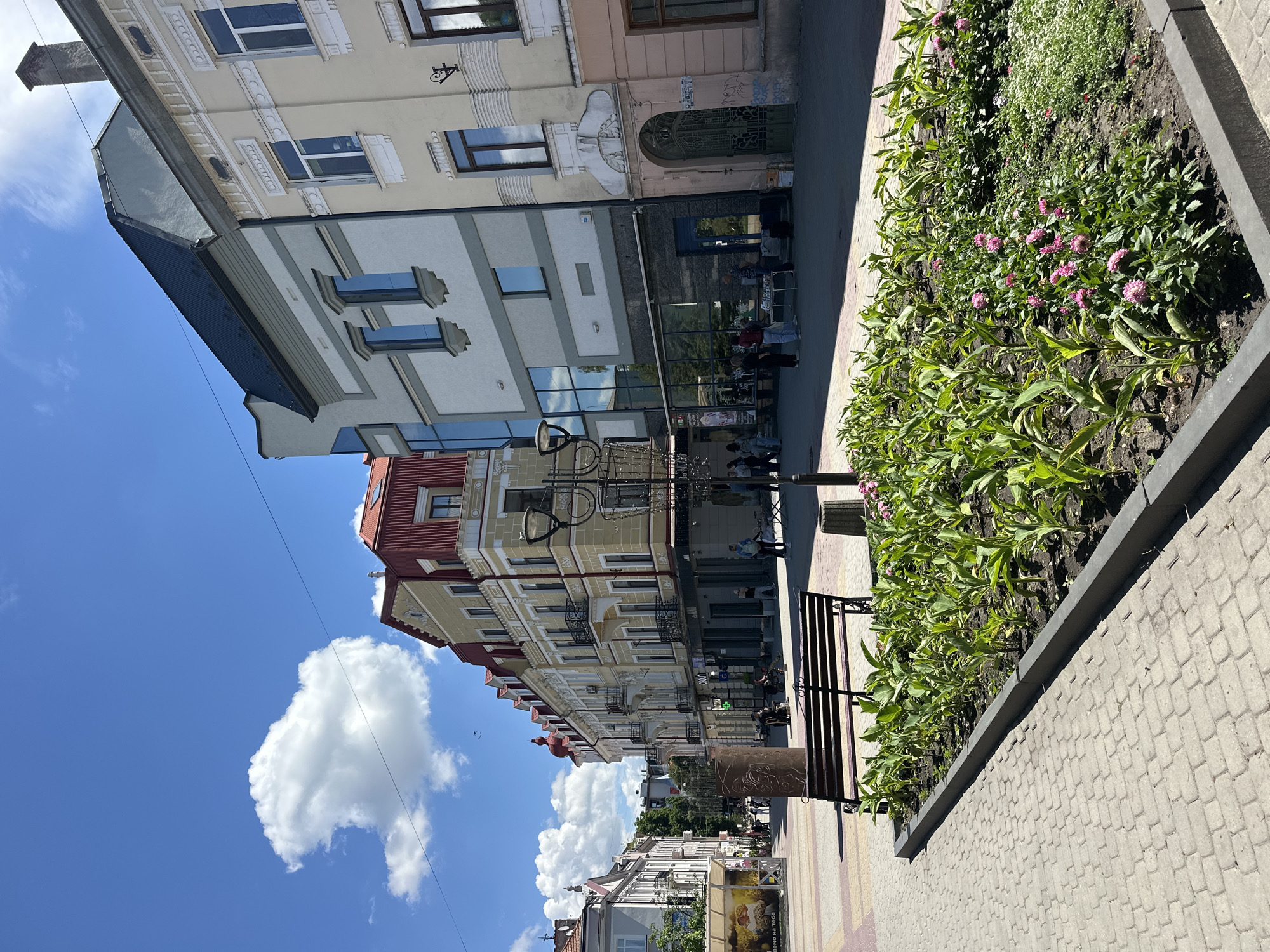 The image depicts a sunlit pedestrian street in Ternopil, Ukraine, likely a city center given the architecture and pedestrian activity. The scene features several multi-story buildings with distinct architectural styles, some adorned with decorative elements and balconies, and others with more modern glass facades.
In the foreground, a paved walkway made of interlocking bricks leads towards the buildings. A dark-colored park bench is situated beside a raised flower bed containing lush green plants and some small pink flowers. Adjacent to the flower bed is a trash receptacle.
Further into the scene, various individuals are visible walking and interacting. Some are browsing at a stall set up on the sidewalk, displaying what appear to be books or small items. Others are walking past or sitting on benches. The sky is a clear blue with scattered white clouds, indicating pleasant weather. A lamppost stands near the flower bed, and a tall, thin sculpture or pillar is located behind the bench. The buildings lining the street have storefronts on the ground floor. The overall impression is of a vibrant urban public space on a bright day.